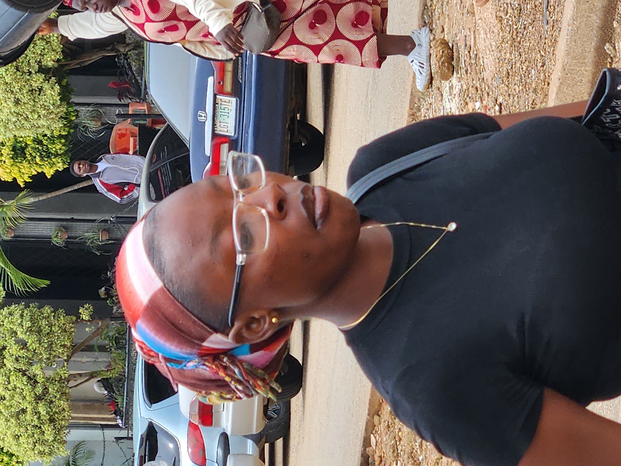 In the city of Unknown, Unknown, a medium shot captures a Black woman wearing glasses and a black t-shirt. She has a gold earring and a gold necklace with a pendant. Her hair is covered by a colorful headscarf. To her right, a person in a white jacket with red and grey stripes stands near a car. Further to the right, a Black woman wearing a patterned dress and white shoes is visible. In the background, there are parked cars, trees, and buildings. The ground is a mix of dirt and gravel.