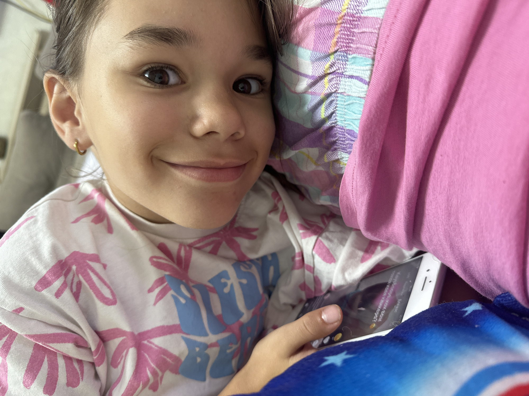 The image shows a close-up of a young person lying down, looking at a smartphone. The person is wearing a white t-shirt with a pink and blue floral pattern. The smartphone is white and displays some text in a foreign language. The person has a faint smile and their eyes are visible. The background is out of focus, but appears to be a domestic setting. There is no indication of the city of Uva, Russia in the image.