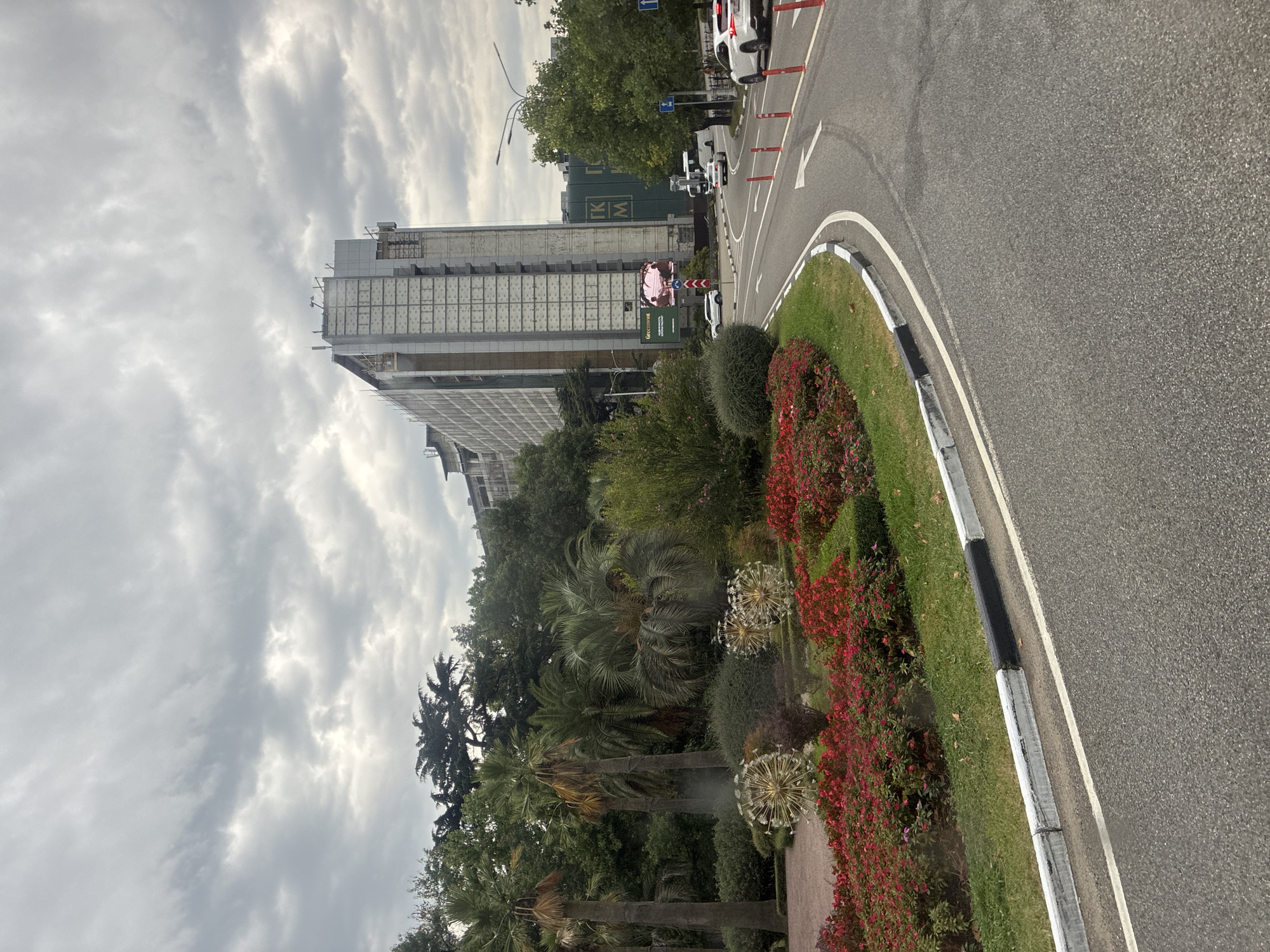 The image depicts a cloudy day in Sochi, Russia, with a wide asphalt road curving through a landscape featuring lush greenery and a large building under construction.
In the foreground, a paved road with white and black painted curbs dominates the lower half of the image. The road has lane markings and several traffic cones. To the left of the road is a neatly manicured garden bed filled with red flowers and green bushes. Behind the garden, a line of palm trees and other lush green trees creates a dense background. Two large, spherical, decorative sculptures resembling dandelions are visible in the garden.
In the midground, a tall, modern building, appearing to be under construction or renovation, rises prominently. Its facade is partially covered with scaffolding and protective netting, revealing sections of grey and white paneling. To the right of this building, a smaller structure with large green panels displaying "GK M" lettering can be seen. Several vehicles, including cars and a white bus, are visible on the road. A billboard advertising "Greenmont" is also present.
The background is filled with a dense canopy of trees, suggesting a park or a well-landscaped urban area. The sky is overcast with heavy grey clouds. The overall scene conveys an urban environment with elements of landscaping and ongoing development.