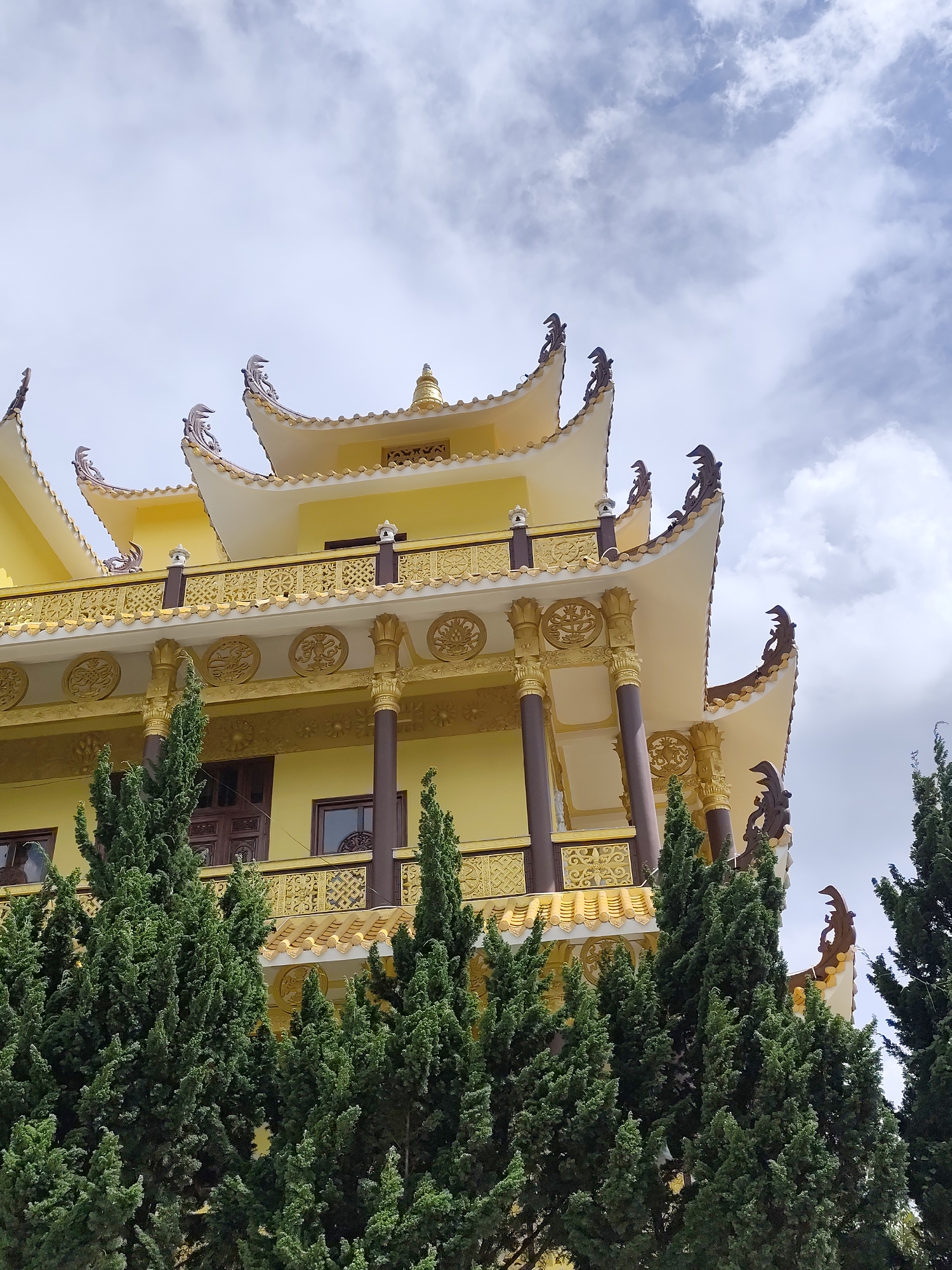 The image captures a multi-tiered, yellow Buddhist temple in Da Lat, Vietnam. The temple's architecture exhibits ornate details with gold accents, layered roofs, and decorative carvings along the edges. Brown columns support the building. Evergreen trees partially obscure the lower portion of the temple. The sky is overcast.