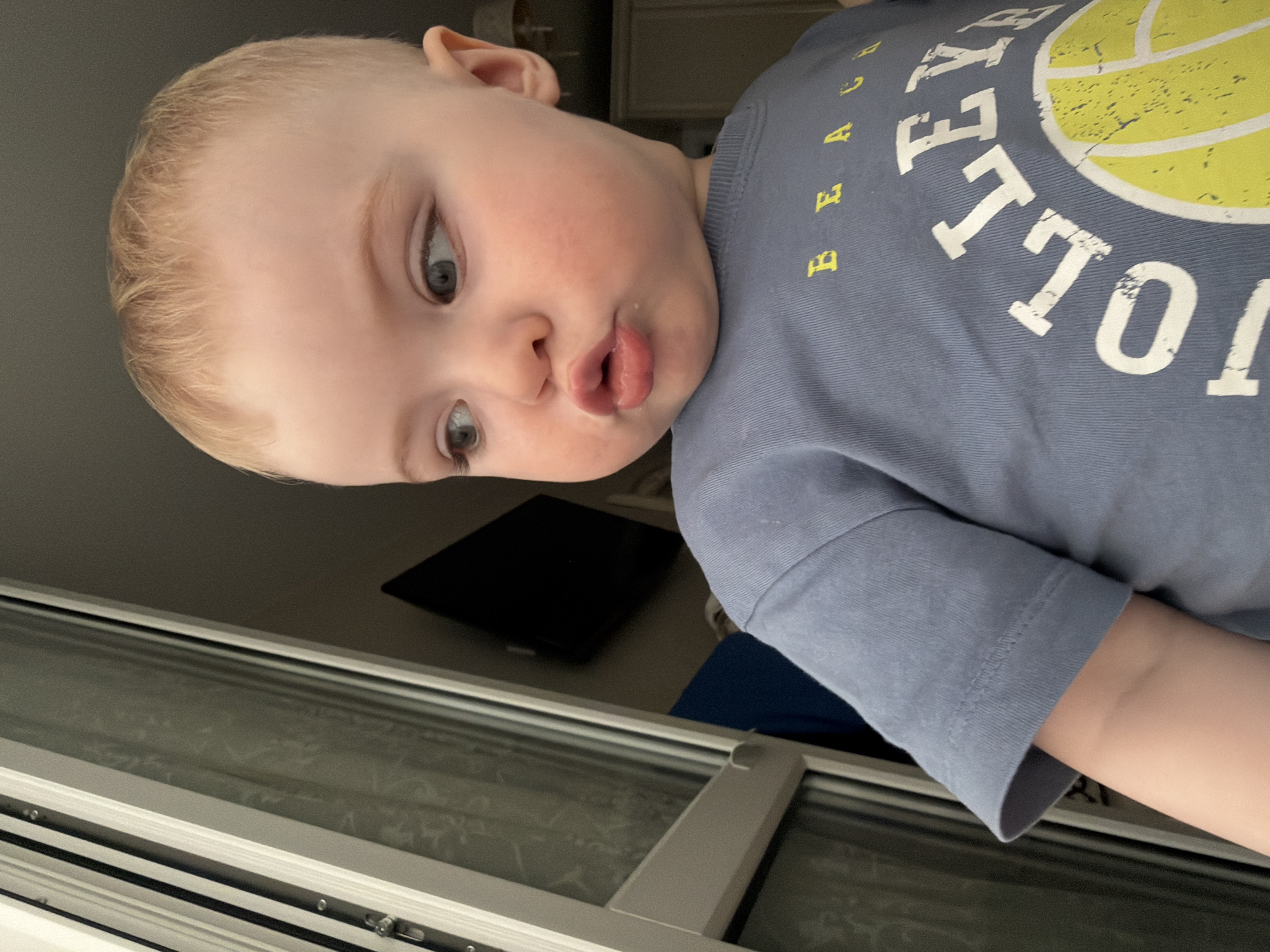 A baby with fair skin, blonde hair, and grey eyes is positioned close to the camera, wearing a blue t-shirt with the word "VOLLEYBALL" printed on it in white and yellow. The baby's face is slightly puckered, and the background includes a window with a frosted glass pane. Behind the window, a dark object, possibly a tablet, is visible. The scene does not provide enough information to determine its specific location in Votkinsk, Russia.