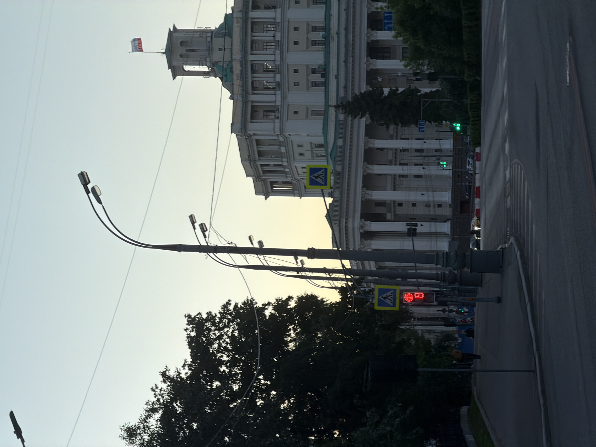 The image depicts a street scene in Moscow, Russia. A multi-story building with classical architectural elements and a Russian flag on the top is in the background. Street lights and power lines intersect the sky. Trees obscure some parts of the scene. A pedestrian crossing sign is visible, along with a traffic light displaying a red signal with the number '16'. A section of a road is also in view. People are on the sidewalk.