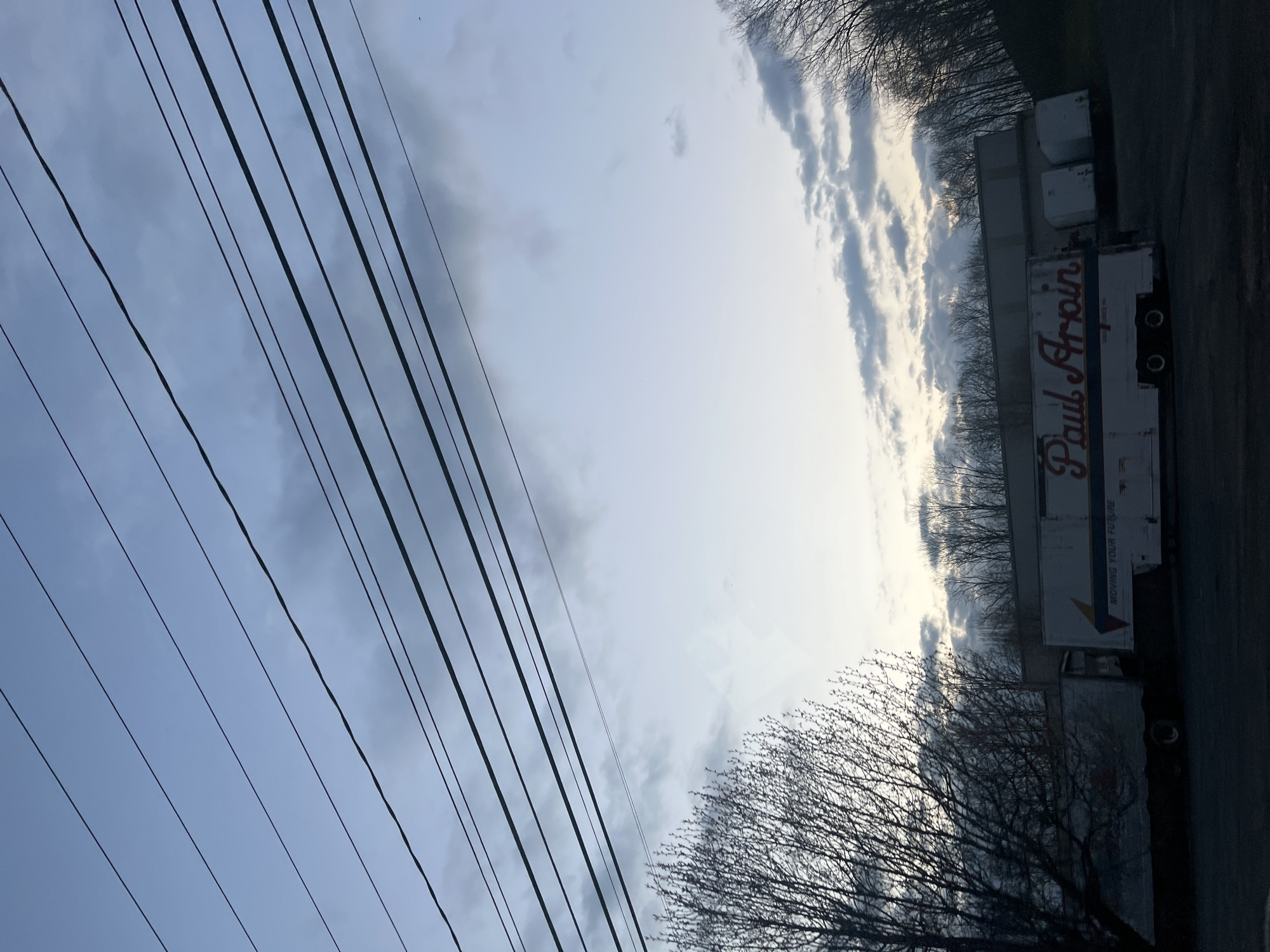 The image shows a row of parked semi-trucks on a street next to a building in Marlboro, United States of America. The trucks have the name "Paul Arpin" printed on their sides. There are bare trees and overhead power lines in the foreground. The sky is cloudy.