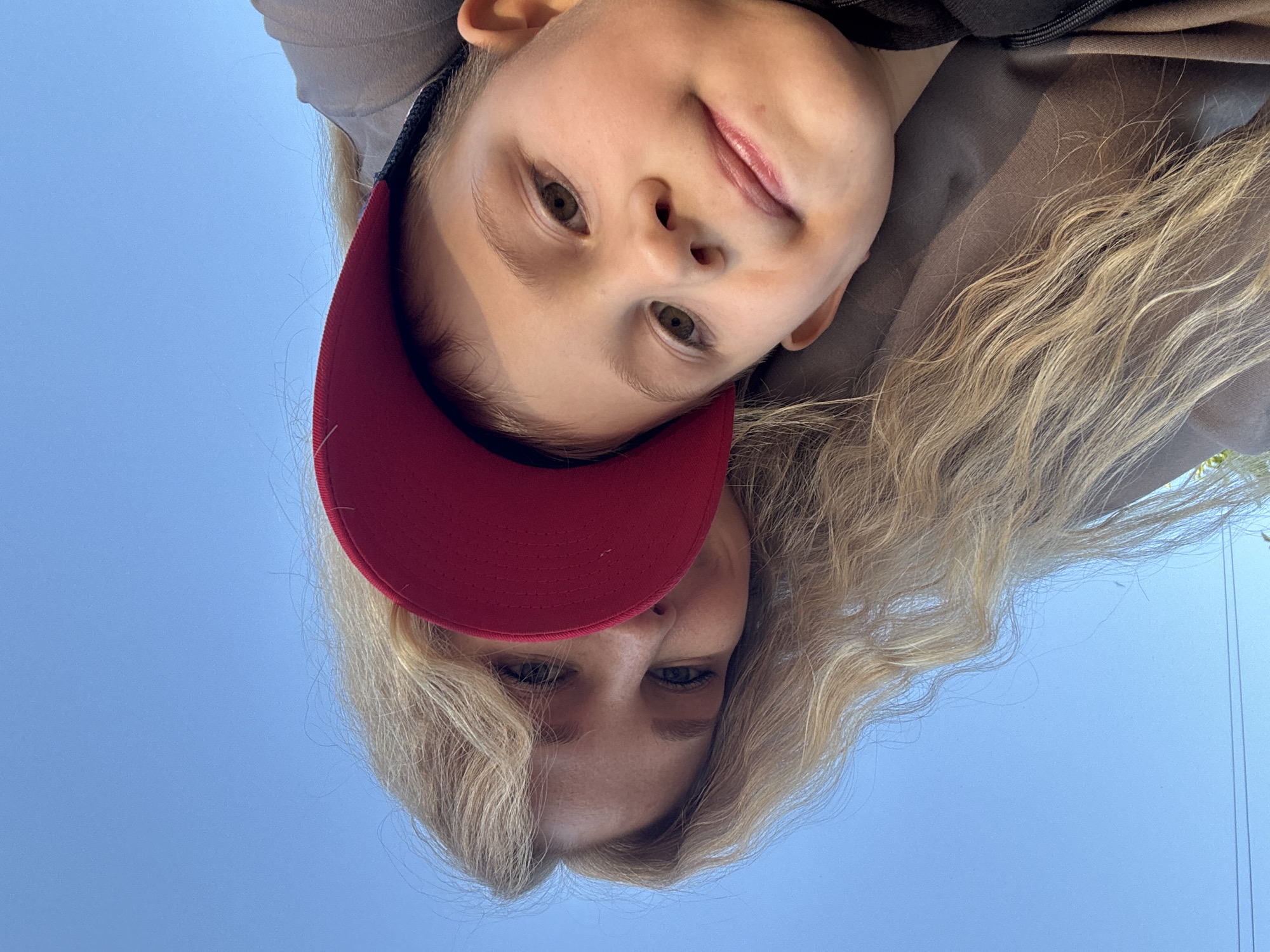 An outdoor scene in Rozhdestveno, Russia, captures an adult female and a young male child from a low, upward-looking perspective against a clear, bright blue sky. The subjects are positioned diagonally across the frame, with the adult female on the left and the child on the right. The female has long, wavy blonde hair and light-colored eyes, with her face partially obscured by the child and a red baseball cap. She is wearing a dark, possibly grey or brown, top. The child, approximately 5-8 years old, wears a prominent red baseball cap with a dark brim and a dark-colored top. Both individuals are looking directly towards the camera. The background is almost entirely open blue sky, with a faint, thin dark line (likely a utility wire) visible in the lower-left quadrant and minimal, dark green foliage barely discernible at the bottom right edge.