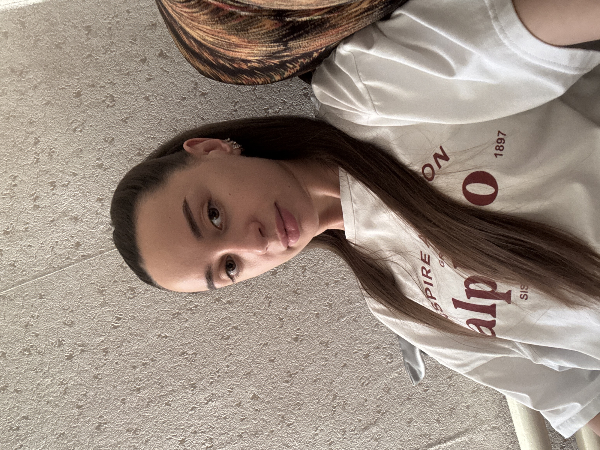 A woman with long brown hair is lying on a textured beige surface. She is wearing a white t-shirt with the word "alpine" and the year "1897" printed on it in red. The top of a brown patterned object is visible in the upper part of the frame. The scene does not obviously appear to be in Goncharka, Russia.