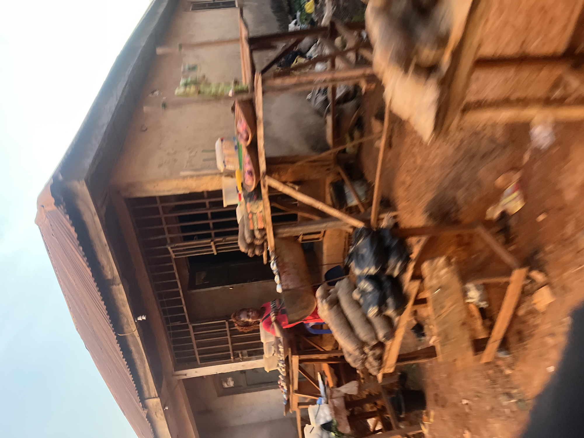 The image shows a two-story building under construction in Abuja, Nigeria. The building's facade is a rough concrete texture. A corrugated metal roof covers the structure. A wooden scaffold surrounds the building, supporting numerous bags of materials, likely cement or sand. A woman wearing a red shirt is visible through the scaffold's framework. The surrounding environment suggests an urban area with dirt ground and signs of construction debris.