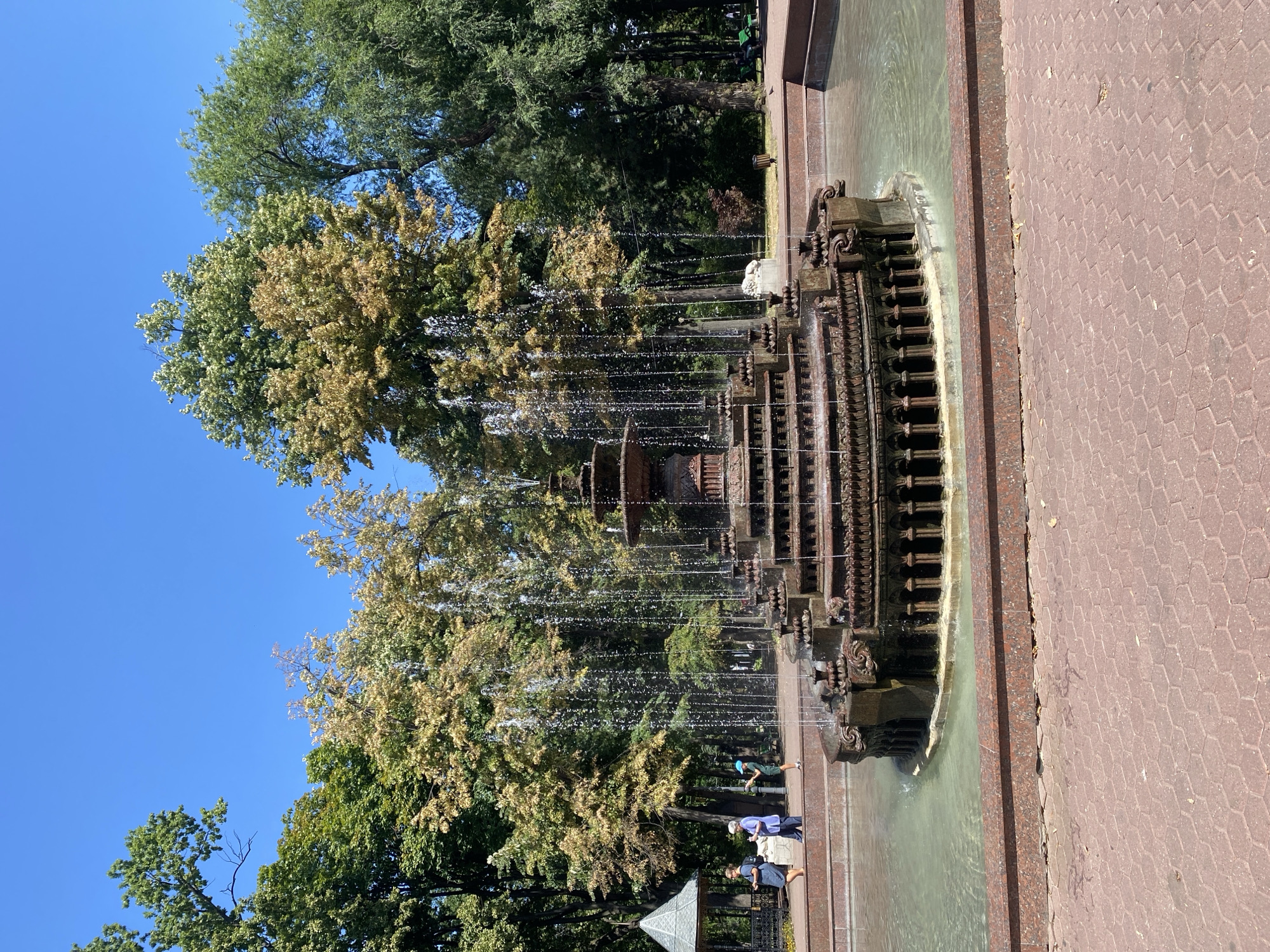 The image shows a large, ornate fountain in the foreground, with water cascading down multiple tiers. The fountain is made of dark metal and has intricate detailing. Behind the fountain, a park setting is visible, with lush green trees and a clear blue sky. Some people are walking in the park, and there is a small gazebo-like structure on the left side of the frame. The ground in the foreground is paved with red hexagonal tiles. This fountain is located in Ștefan cel Mare Central Park in Chișinău, Moldova.