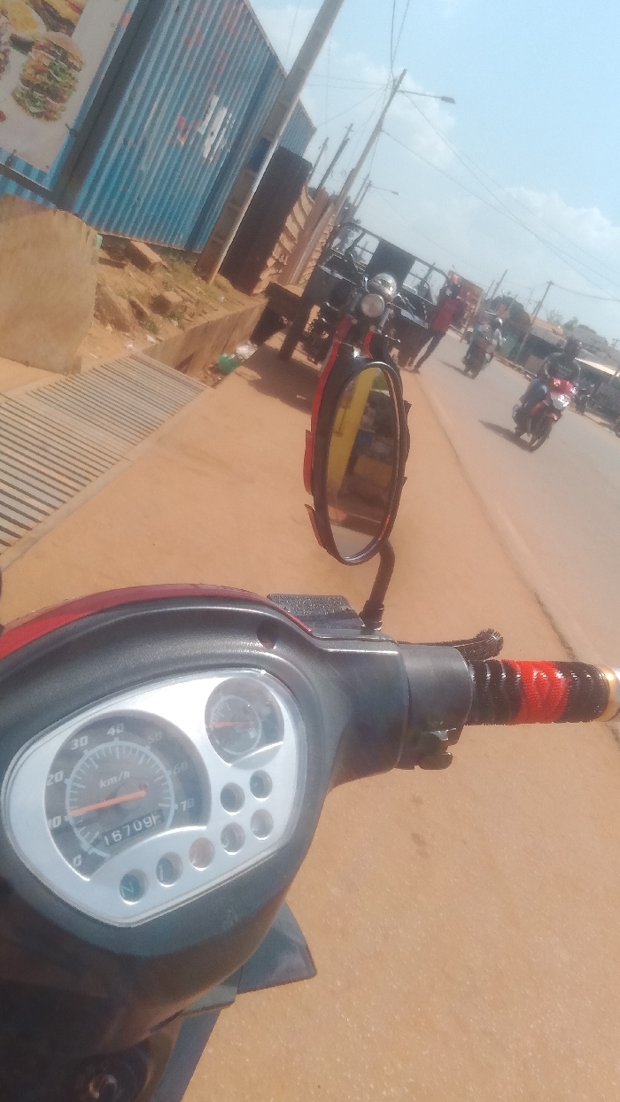 The image depicts a bright daylight scene on a dusty street in Korhogo, Côte d'Ivoire, viewed from the dashboard perspective of a stationary or slow-moving red and black scooter.
In the immediate foreground, the scooter's dark grey and black plastic dashboard is prominent, featuring a silver-colored instrument cluster. This cluster includes a speedometer showing a range up to 70 km/h with an orange needle at zero, an odometer displaying "16709f," a smaller circular fuel gauge indicating a half-full tank, and several indicator lights. The right handlebar is visible with a distinctive black and red woven grip. A red and black rear-view mirror reflects a blurred portion of the street.
To the left of the scooter, a light brown concrete sidewalk or curb is visible, along with a metal drainage grate. Beyond the curb, the ground is dusty, leading to a large, corrugated blue structure, resembling a modified shipping container, which appears to be a commercial establishment. A sign affixed to its left displays a large image of a hamburger and some obscured white text.
The main street, composed of light brown, unpaved or dust-covered ground, stretches into the distance. Utility poles with overhead wires line the street on both sides. On the right side of the road, a three-wheeled cargo motorcycle (tricycle motor) is either parked or moving slowly away from the camera. Behind it, a person wearing a red and black patterned shirt is visible. Further down the road, several other motorcycles are present, some with riders, including one person on a red motorcycle. The sky is clear and light blue with scattered white clouds. The overall environment suggests an active urban or semi-urban setting under sunny conditions.