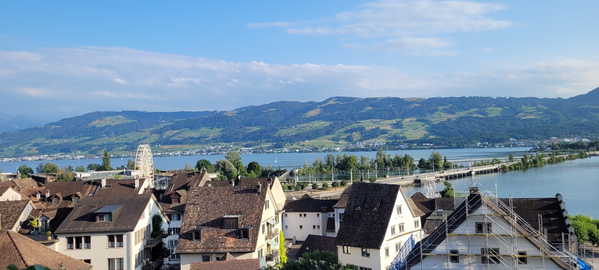 The image shows a panoramic view of the city of Rapperswil-Jona, Switzerland, on a clear day with a blue sky and scattered clouds. In the foreground, traditional Swiss architecture with brown tiled roofs dominates the scene. Several multi-story buildings are visible, some with dormer windows and white facades. One building in the foreground appears to be under renovation, with scaffolding surrounding it.
Beyond the buildings, a body of water, likely Lake Zurich, stretches into the distance. A long bridge or causeway crosses the water, connecting different parts of the landscape. Trees and greenery are interspersed along the waterfront and on the distant hills.
The background is characterized by rolling hills and mountains covered in lush green vegetation. Small settlements and scattered houses are visible on the slopes of these hills, indicating a rural or semi-rural environment. A Ferris wheel can be seen to the left of the image, suggesting a recreational area or amusement park. The overall impression is a scenic and tranquil landscape, typical of Swiss lakeside towns. No people are clearly discernible in the image.