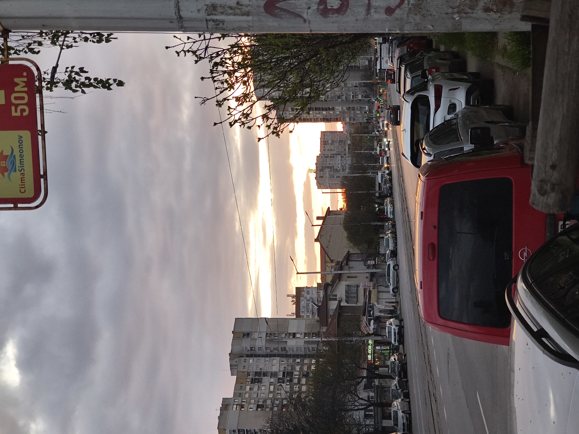 The image displays a city street scene, rotated 90 degrees counter-clockwise, capturing a moment at either sunrise or sunset. The sky, a prominent feature, showcases a dramatic blend of grey clouds and vibrant orange-yellow light from the low sun, casting a warm glow across the urban environment.
A multi-lane road stretches into the distance, with numerous cars parked along its right side. In the immediate foreground, a red van, identifiable by an Opel logo on its rear, is parked next to a white SUV. Further along the street, various other vehicles, including cars and vans, are visible, some in traffic.
The street is flanked by multi-story buildings, characteristic of residential or mixed-use structures in Eastern European cities, with some appearing as block-style apartments. Trees with sparse, budding leaves line the roadside, suggesting an early spring or late autumn setting.
In the far left foreground, a red and yellow sign hangs from an overhead structure. It clearly displays "50M." at the top and "ClimaSimeonov" with a logo featuring a house and a wave, likely advertising an air conditioning or climate control business. A distant shopfront sign bears Cyrillic text: "ХРАНА • НАПИТКИ • ЦВЕТЯ," which translates to "FOOD • DRINKS • FLOWERS." This Cyrillic script, along with the architectural style, confirms the location as Sofia, Bulgaria. The scene captures a tranquil urban moment at dawn or dusk, devoid of specific events or prominently featured people.