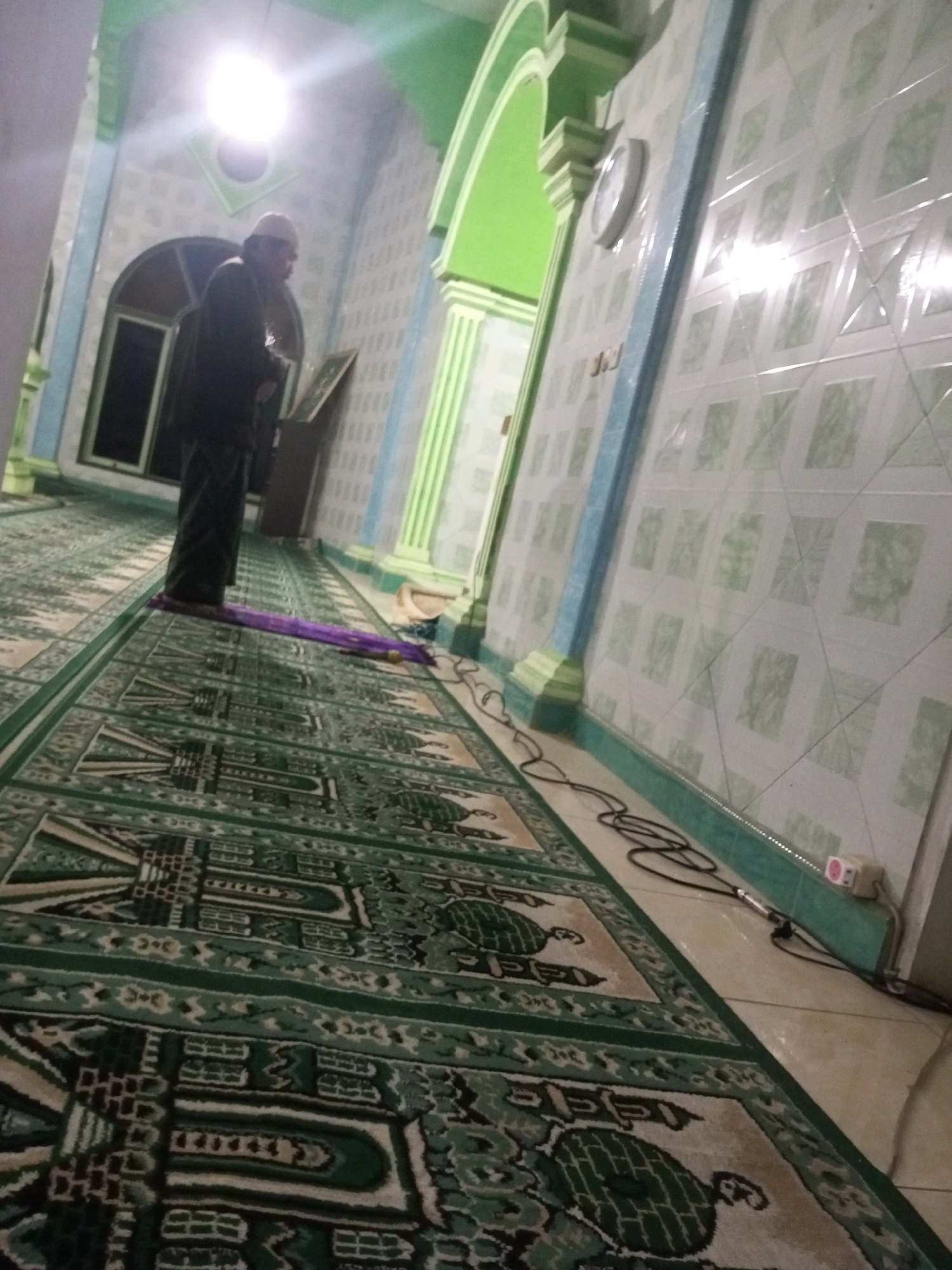 A man in traditional attire, including a cap, stands on a purple prayer rug in what appears to be a mosque. He is facing away from the camera and slightly to the left, with his hands by his sides. The floor is covered with a green carpet featuring intricate geometric patterns. In the background, there is a wall covered in patterned tiles, and a clock is visible on the wall. The lighting suggests an indoor environment, possibly in the city of Andongsili, Indonesia.