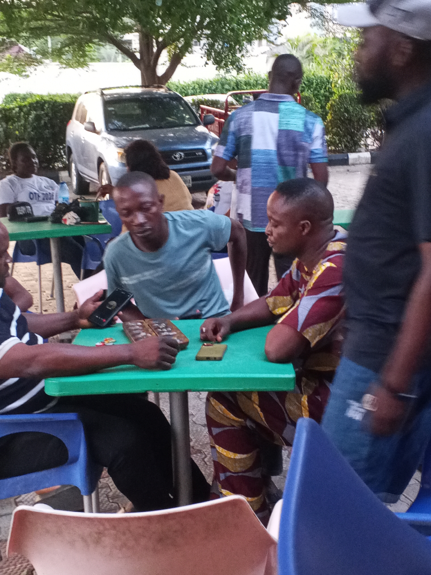 In Lokoja, Nigeria, a group of men are gathered outdoors at tables with green tops and blue chairs. One table has three men playing a game with a wooden board and small pieces. One man holds a cell phone. Two other men are at another table nearby. A silver SUV is parked in the background, partially obscured by trees and greenery. Another man stands nearby.