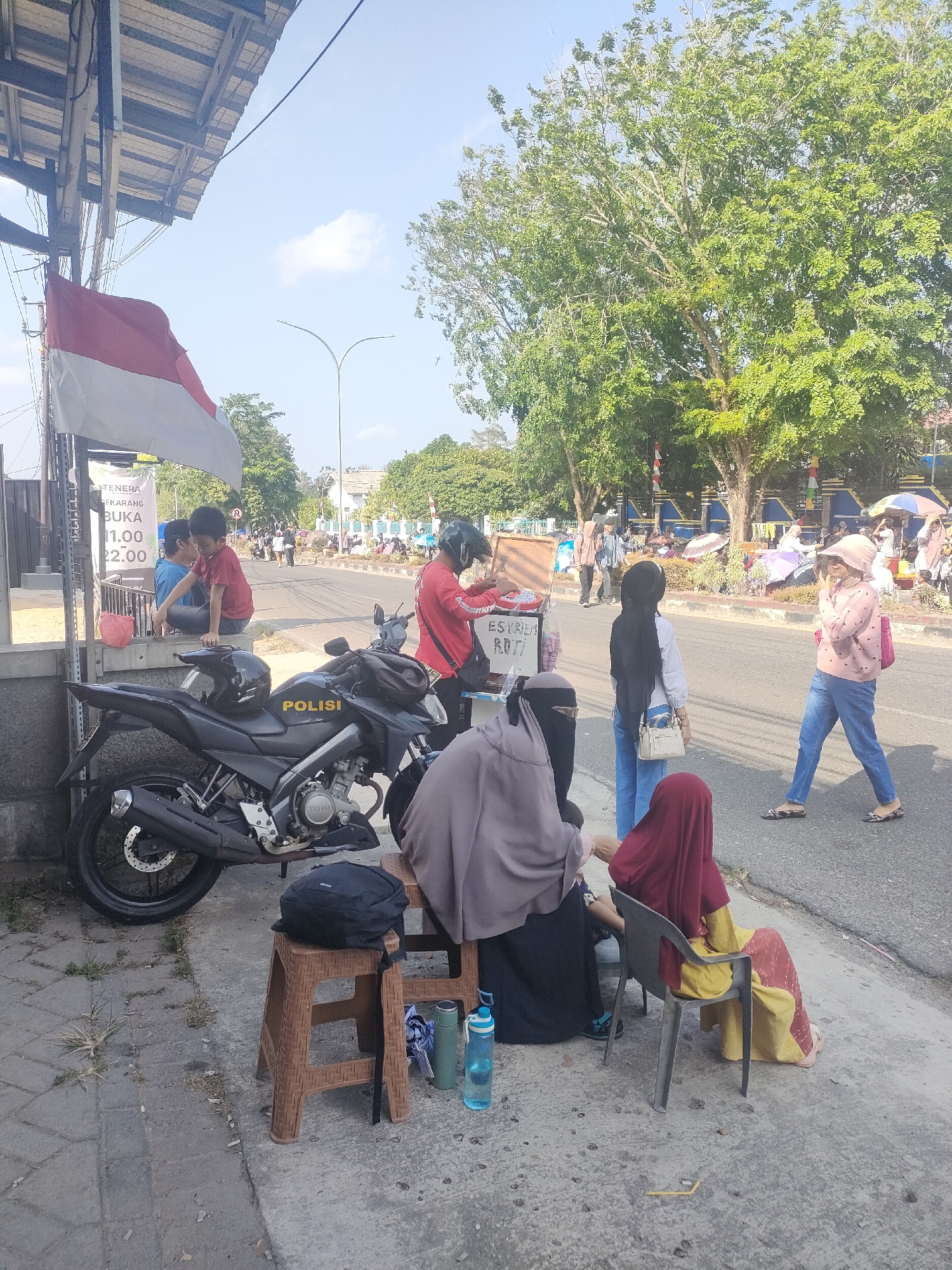 The image captures a street scene in Sungailiat, Indonesia, on a bright, sunny day. The foreground features a black police motorcycle with the word "POLISI" clearly visible on its side. A black backpack rests on a brown plastic stool next to the motorcycle, with two water bottles and a thermos nearby. Several individuals are present. In the immediate foreground, a person wearing a red hijab and a yellow and red patterned dress is seated on a black plastic chair. To their left, partially visible, another person is wearing a grey hijab and black clothing. On the left side of the image, a child in a red shirt is sitting on a concrete ledge, with another person in a blue shirt beside them. A red and white Indonesian flag flies from a pole in the background. Further back, a food cart with "ESCREAM Roti" visible on it is being attended by a person in a red shirt and helmet. Other people, some carrying umbrellas, are visible along the street and sidewalk, suggesting a public gathering or market. Trees line the far side of the road, and buildings and a blue fence can be seen in the distance. The overall atmosphere appears to be casual and active, with a mix of everyday life and what might be a local event or gathering.
