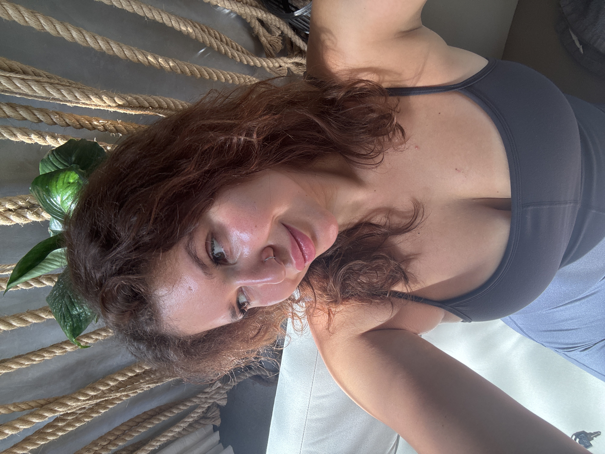 A woman with brown hair is posing for a close-up, angled selfie. She is wearing a gray top and appears to be indoors. In the background, there are thick ropes suspended, and a green plant is visible to the left. Based on the prompt, the location is likely in the Ubud District, Indonesia, though this cannot be confirmed from the image alone.