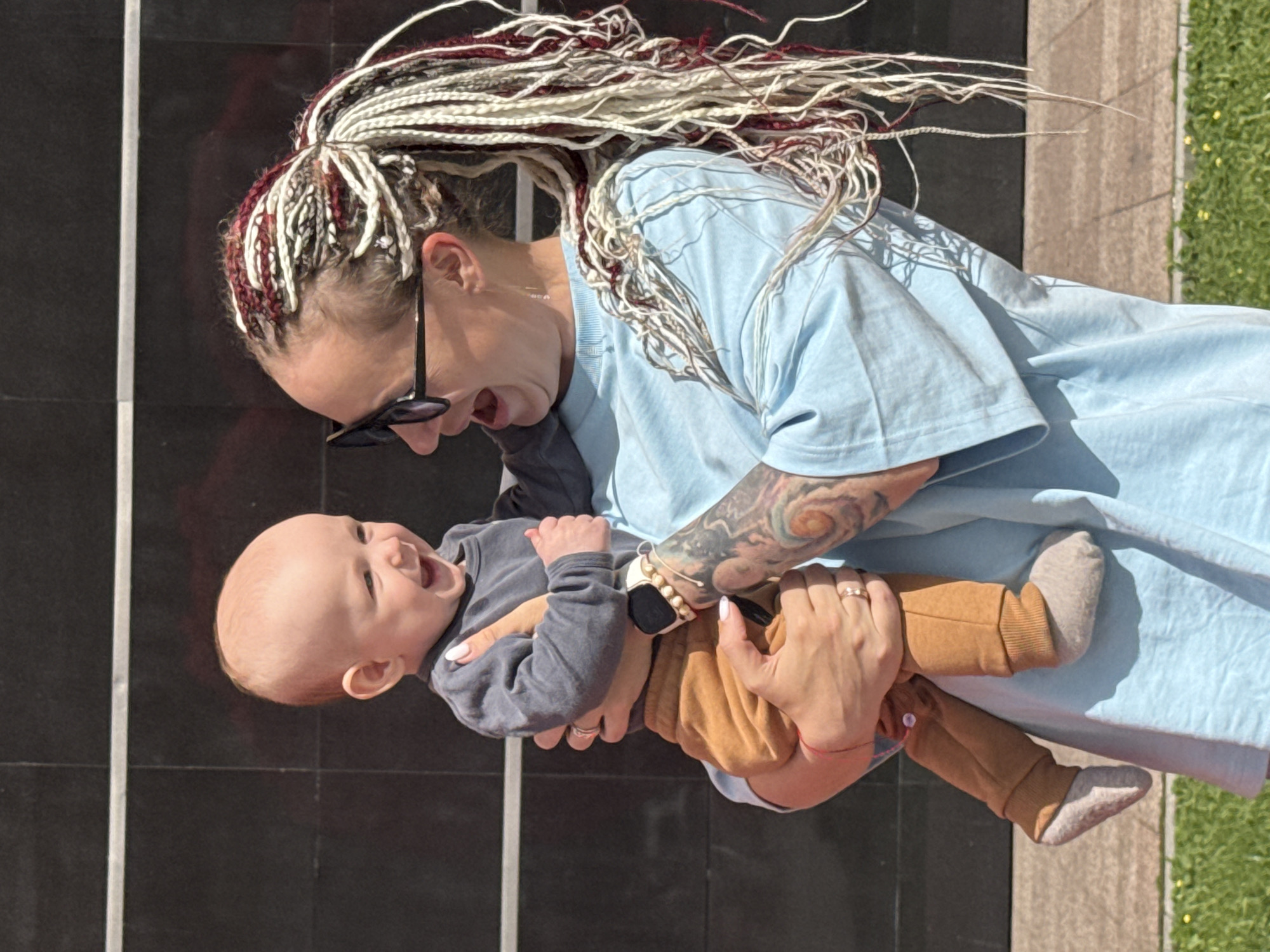 The image features a woman with long, braided hair in white and burgundy, wearing a light blue t-shirt and sunglasses. She is holding a baby who is wearing a grey long-sleeved shirt and brown pants with grey socks. The woman has tattoos on her arm and is wearing rings on her fingers. The baby appears to be laughing and looking upwards.
They are outdoors, with a dark, tiled wall behind them and a patch of green grass with yellow flowers to the right. The lighting suggests it is daytime, likely sunny. The scene appears to be a casual, familial moment. The woman's expression is one of joy and affection as she interacts with the baby. The overall mood is happy and heartwarming. There are no visible text elements in the image. The provided location context is Moscow, Russia.