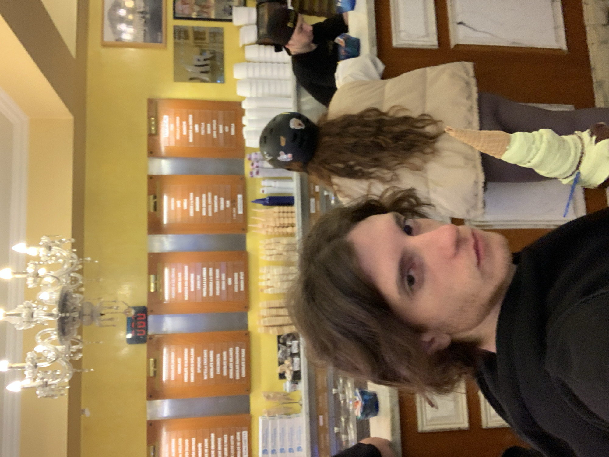 The image depicts a brightly lit ice cream shop in Buenos Aires, Argentina, recognizable by the Spanish-language menu boards displaying flavors and prices. Three people are visible. A person in the foreground is taking a selfie. Another person in the middleground is wearing a helmet, holding an ice cream cone. A person is standing behind the counter, presumably serving customers. The shop has a classic interior design, featuring a chandelier, yellow walls, and framed pictures and signage. Stacks of disposable cups are visible behind the counter.