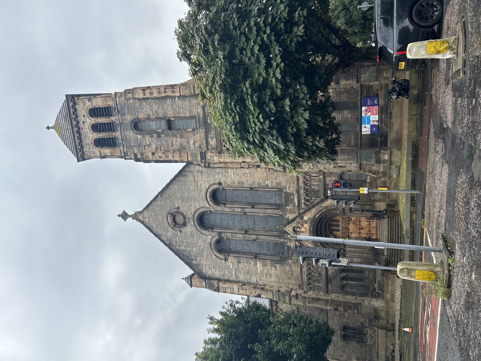 The image shows the exterior of a stone church in the city of Edinburgh, United Kingdom, under an overcast sky. The church features a prominent bell tower with arched openings and a cross at its apex. The main facade of the church has large stained-glass windows and an ornate wooden double door at the entrance, accessed by a flight of steps. A large, leafy tree partially obscures the right side of the church. In the foreground, there are road markings, traffic lights, and street furniture, including yellow bollards. A black SUV and a motorcycle are parked near the church. The ground appears wet, suggesting it may have recently rained. The overall impression is of a historic religious building within an urban environment.