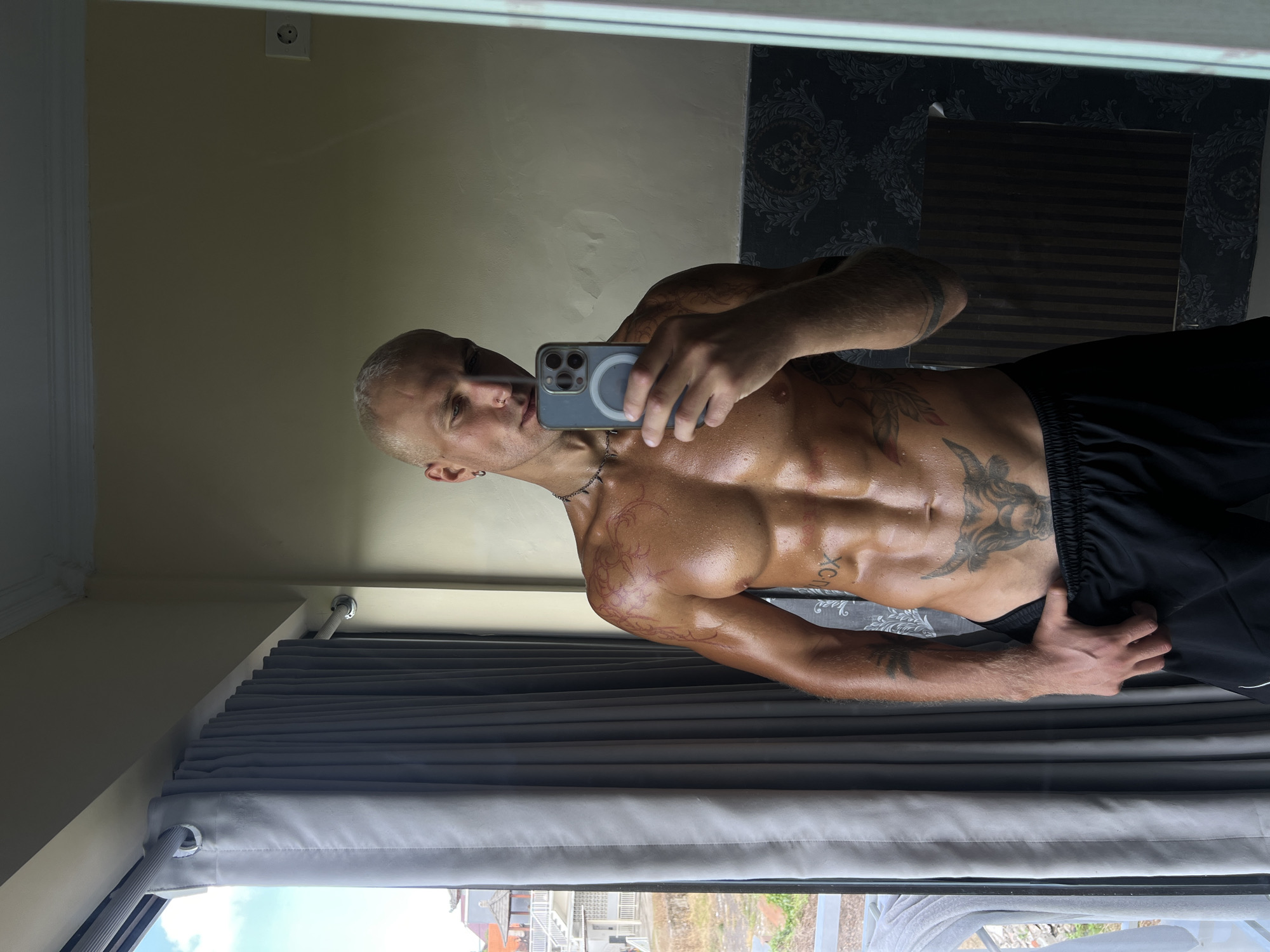 The media file is a selfie taken by a man with visible tattoos on his chest, arm, and shoulder. He is shirtless, revealing his muscular physique, and is wearing black shorts. The man is holding a smartphone in his right hand, capturing the reflection in a mirror.
The setting appears to be an indoor space with a cream-colored wall and a dark patterned wallpaper visible in the mirror's reflection. There are grey curtains in the foreground, partially obscuring a view of what looks like an outdoor scene with buildings and greenery, suggesting a location in North Kuta, Indonesia. The lighting indicates it is daytime. The man's expression is neutral. The image is oriented vertically.