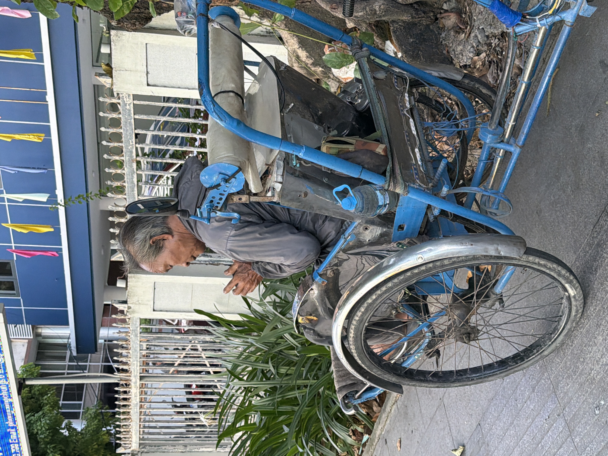 A weathered, elderly Vietnamese man sits in a blue-painted cyclerickshaw parked on a sidewalk. He is wearing a gray jacket and appears to be praying or meditating with his hands clasped together in front of him. The cyclerickshaw is parked next to a large, leafy green plant and a tree with rough bark. Behind the plant and the man is a white metal fence, and beyond that, a blue building with a white stripe and several flags hanging from it. The building appears to be a commercial or municipal establishment. The sidewalk is paved with grey square tiles. The scene suggests a typical street view in a Vietnamese city, likely Nha Trang, given the architectural style and the presence of a cyclerickshaw.