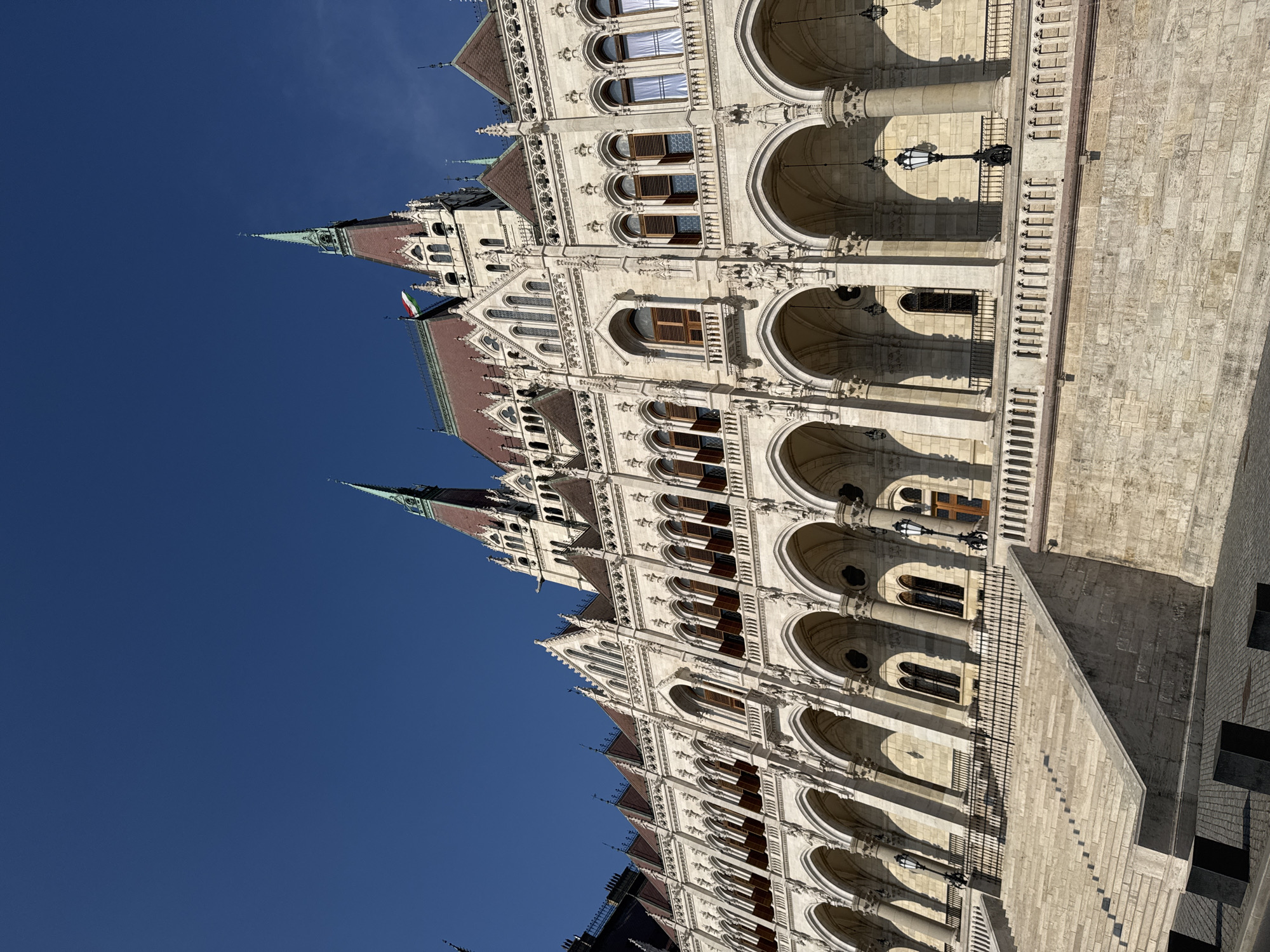This image shows a low-angle, close-up view of the Parliament Building in Budapest, Hungary. The ornate Gothic Revival architecture is evident with its numerous spires, arched windows, and detailed stonework. The building is constructed of light-colored stone, and its red-tiled roof is visible. A Hungarian flag is flying from one of the turrets. The building is bathed in bright sunlight, casting strong shadows, suggesting it is midday on a clear, sunny day. The deep blue sky fills the upper portion of the image. The perspective emphasizes the grandeur and scale of the building, with a section of stone steps and a walkway leading up to the main structure. The scene evokes a sense of history and architectural splendor. No people are visible in the frame.