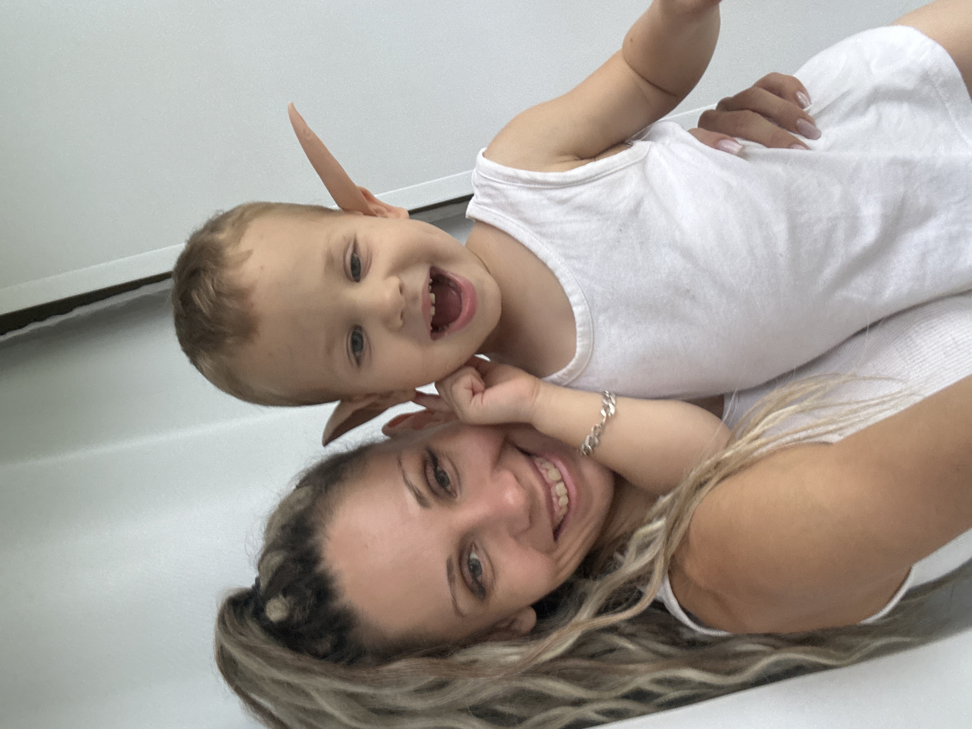 A woman and a young child are posed for a selfie. The woman, on the left, has long, blonde, braided hair pulled into a high ponytail. She is wearing a white tank top and a silver bracelet on her left wrist. Her left hand is supporting the child's head, and her right arm is around the child's torso. The child, on the right, is wearing a white tank top and has large, pointed, prosthetic ears attached to the sides of his head. He is looking at the camera with his mouth open and appears to be smiling or laughing. The background is a plain, light-colored wall, likely indoors, with a thin vertical line, possibly a door frame or the edge of a wall. The image has not been geolocated to Rusanovo, Russia, or any other specific city.