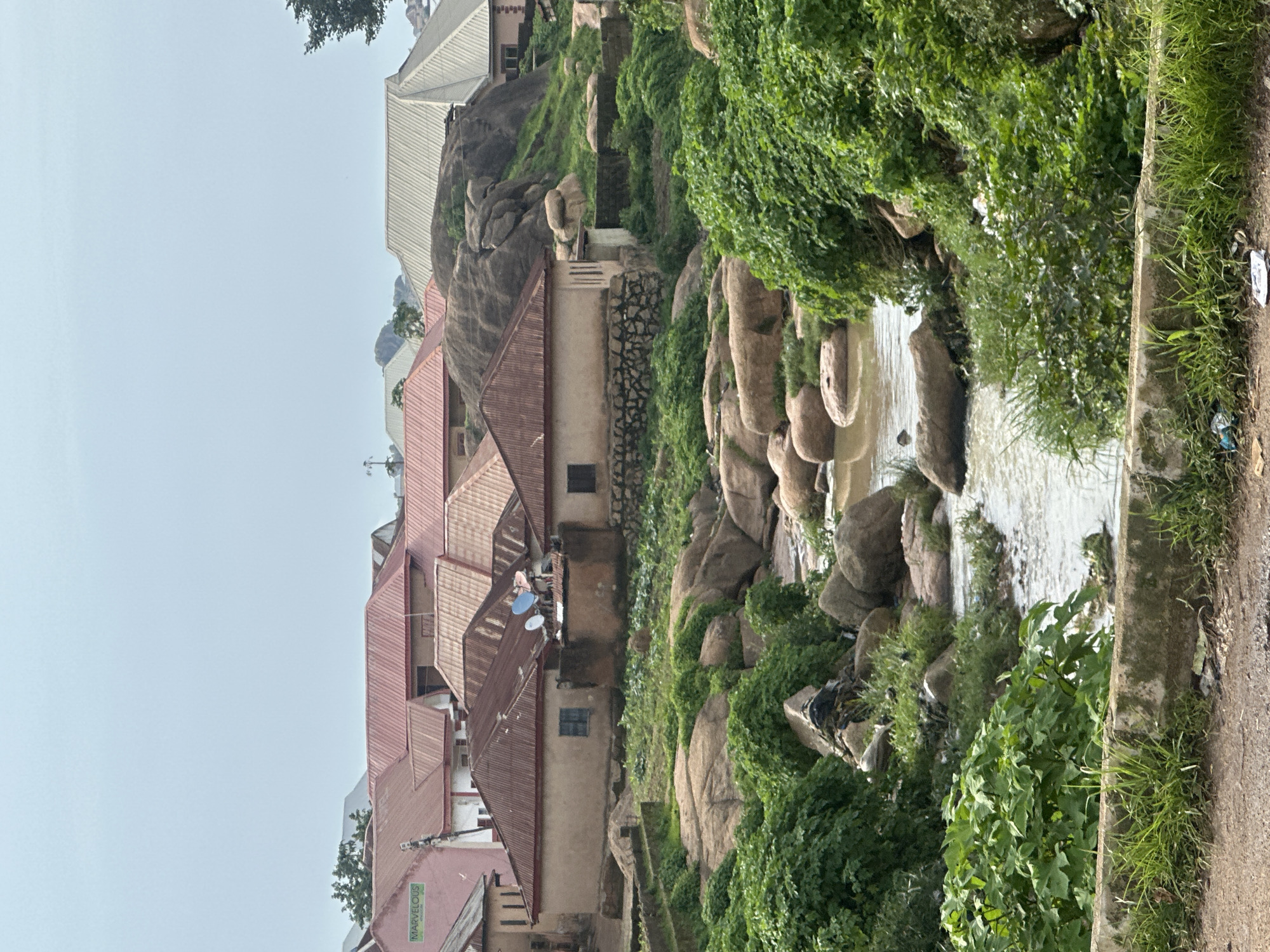 This image captures a scene in Jos, Nigeria, showcasing a natural landscape integrated with residential structures. In the foreground, a rough concrete barrier borders a dirt path, with some scattered litter visible. Beyond the barrier, lush green vegetation and large, weathered boulders dominate the terrain. A narrow, murky stream flows through the rocky landscape, with its banks densely overgrown with various plants.
Further into the scene, several houses with corrugated metal roofs are nestled amongst the rocks and vegetation. The roofs exhibit various shades of red and silver. One prominent building, painted pink and bearing a green sign that reads "MARVELOUS," is visible on the left. The houses appear to be single-story or two-story structures with simple architectural designs. Satellite dishes are mounted on some of the roofs. The background reveals more houses and the undulating terrain characteristic of Jos. The sky is overcast with a pale blue hue. There are no people or animals visible in this frame.