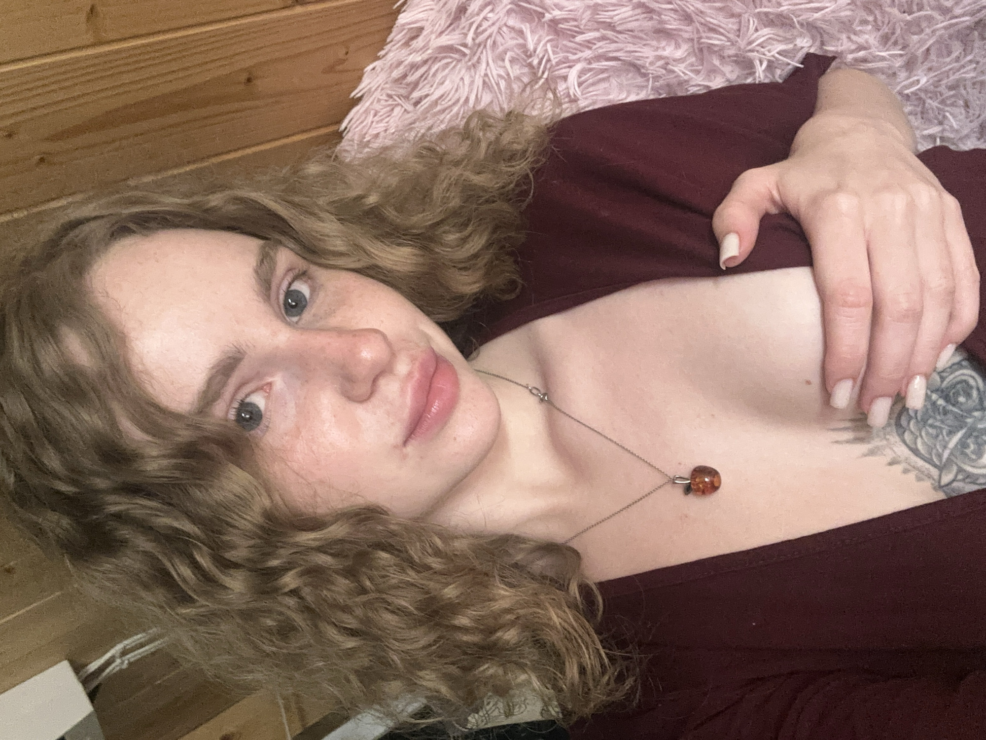 A fair-skinned woman with curly blonde hair lies on her side, facing the camera. She has freckles and blue eyes. She is wearing a maroon-colored top and a silver necklace with a round amber pendant. Her right hand is visible near her chest, with manicured fingernails. A tattoo is visible on her chest, near her right shoulder. The background includes a wooden wall and a pink fluffy surface, potentially a blanket or pillow. No information in the image indicates the location is Vasilyevo, Russia.