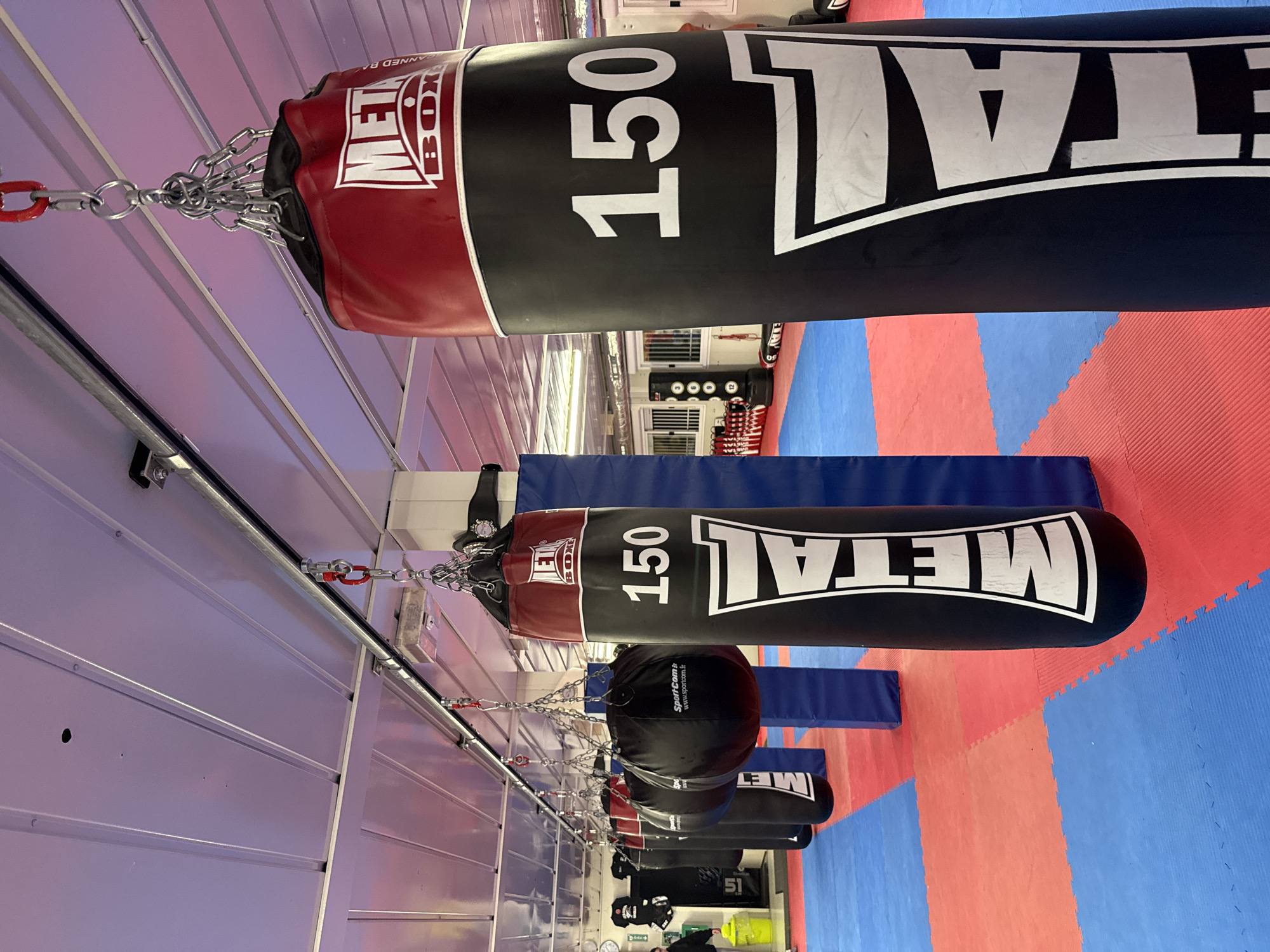 This indoor shot shows a row of black punching bags suspended from the ceiling in a gym. The bags are cylindrical and labeled with "METAL BOXE" in white lettering, along with the number "150". They hang from chains that are attached to the ceiling. The floor is covered in blue and red interlocking foam mats. In the background, there is gym equipment. The location is likely a gym or training facility in Sarcelles, France.