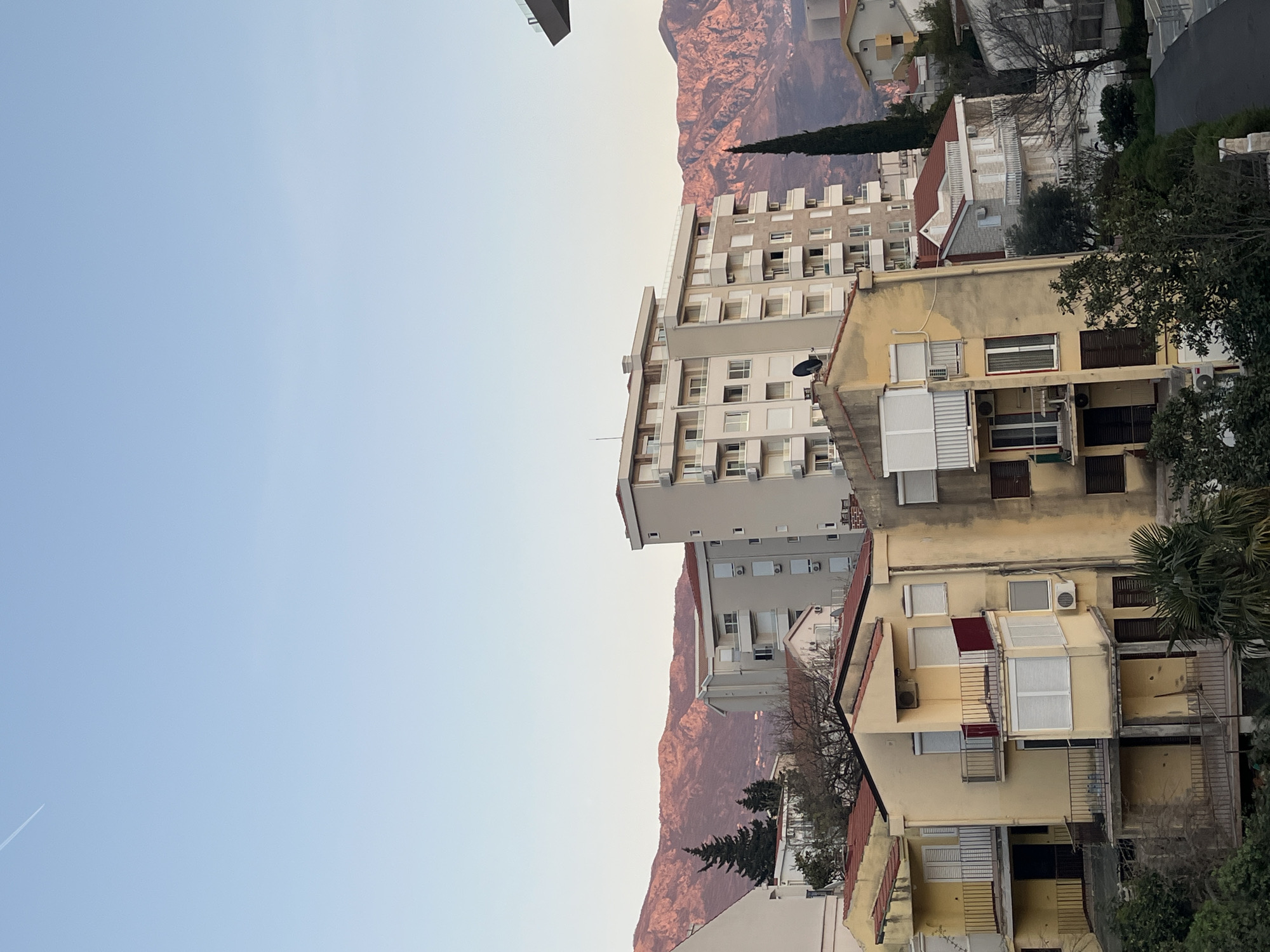 An eye-level outdoor shot depicts a cluster of buildings against a mountain backdrop in Boreti, Montenegro. The building closest to the viewer is a 3-story structure painted in shades of yellow and brown. It features rectangular windows with white-slatted blinds. A metal staircase is visible on the right side of the building. Behind this building, a taller, 8-story apartment block with white balconies and windows rises into the sky. It is light grey in color. Mountains in hues of pink and brown are visible in the background. The sky is a pale blue. At the upper left is a portion of another building.