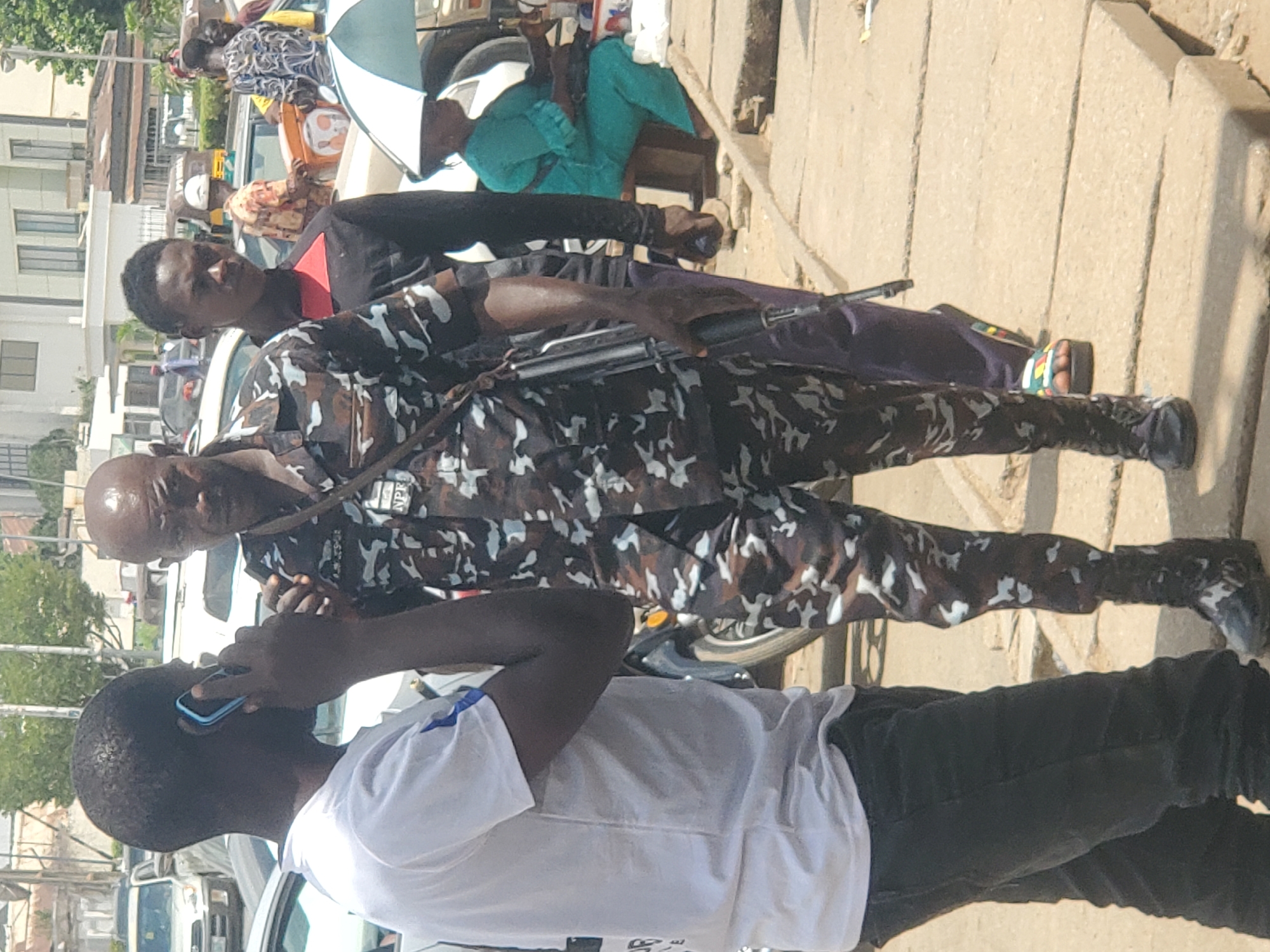 A daytime scene in Jos, Nigeria shows three men on a sidewalk along a street. A man in a white t-shirt is holding a blue-cased mobile phone to his ear, presumably in conversation. Next to him stands a bald man wearing camouflage police uniform, holding a rifle. Behind the policeman, another man in a black and red shirt and purple trousers is looking in the camera's direction. Pedestrians and cars are visible in the background.