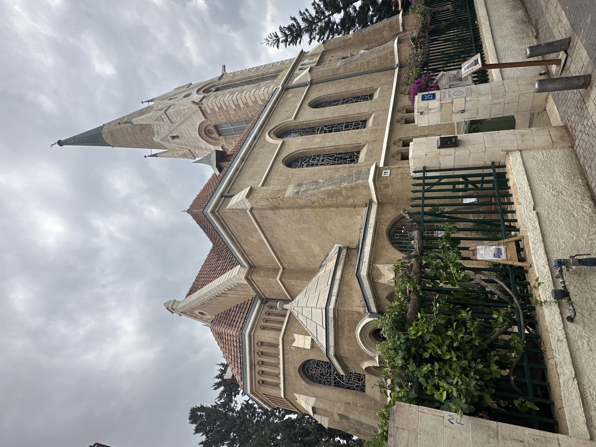 The image shows the Immanuel Church, located in the American-German Colony of Tel Aviv, Israel. The church is a sandstone building with a tall, pointed steeple. The sky is overcast and gray. The church is surrounded by a fence. There are bushes and trees.