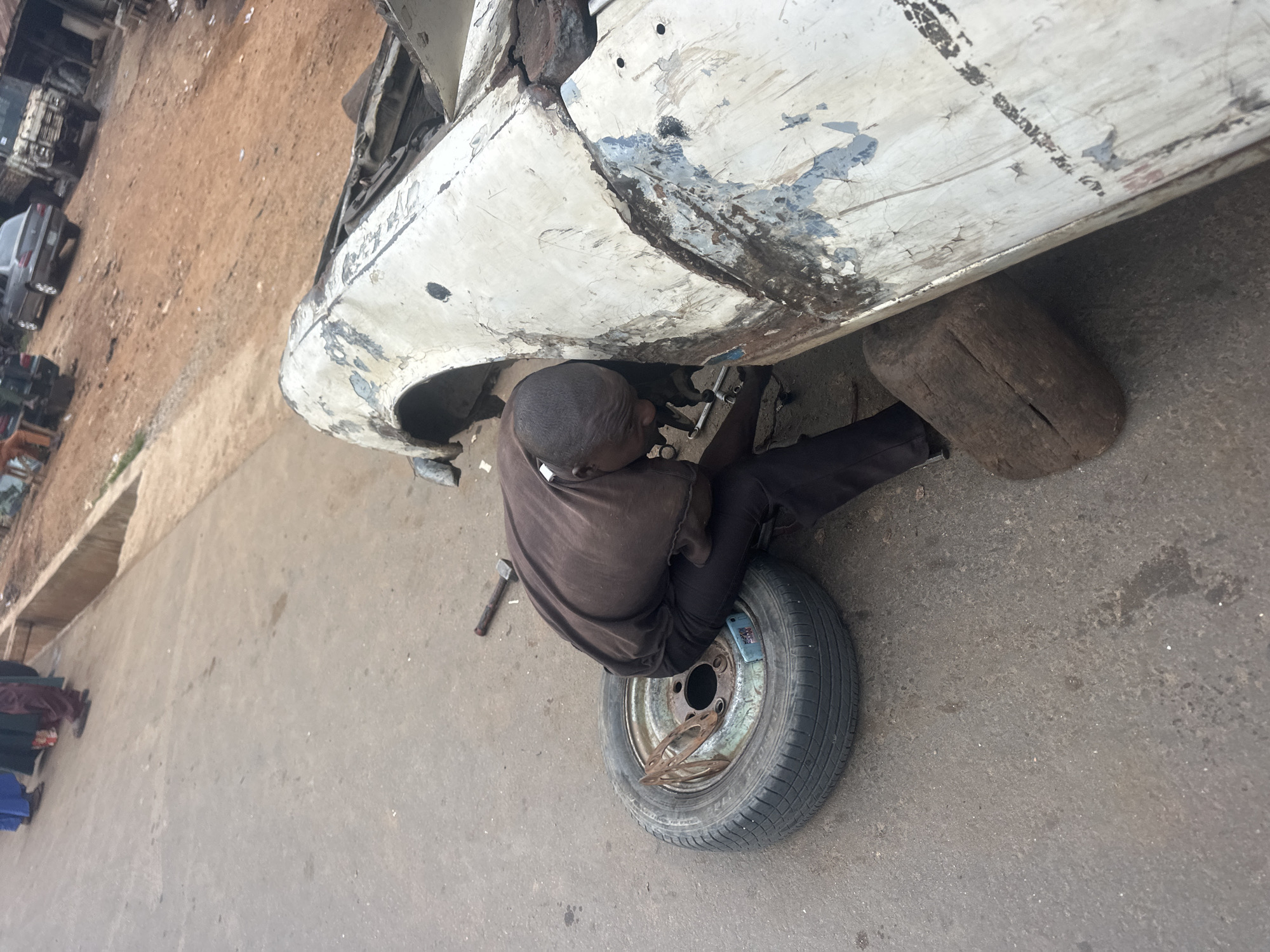 A person is working on a car's wheel. The scene appears to be an outdoor mechanic shop or junkyard, with the dusty ground and debris suggesting a rural or industrial setting. The car itself looks old and heavily weathered, with peeling paint and signs of rust. The person is positioned underneath the car, lying on their back on the ground, working on the wheel which has been removed. They are wearing dark clothing. Tools, including a hammer and some metal parts, are scattered around the wheel. A large, rough-hewn log is used to prop up the car. In the background, other damaged vehicles and piles of scrap metal are visible, reinforcing the impression of a junkyard. The lighting suggests it is daytime, but the weather is not clearly discernible. There is no visible text in the image. The overall impression is one of diligent work in a challenging environment, perhaps indicating a repair or dismantling process.