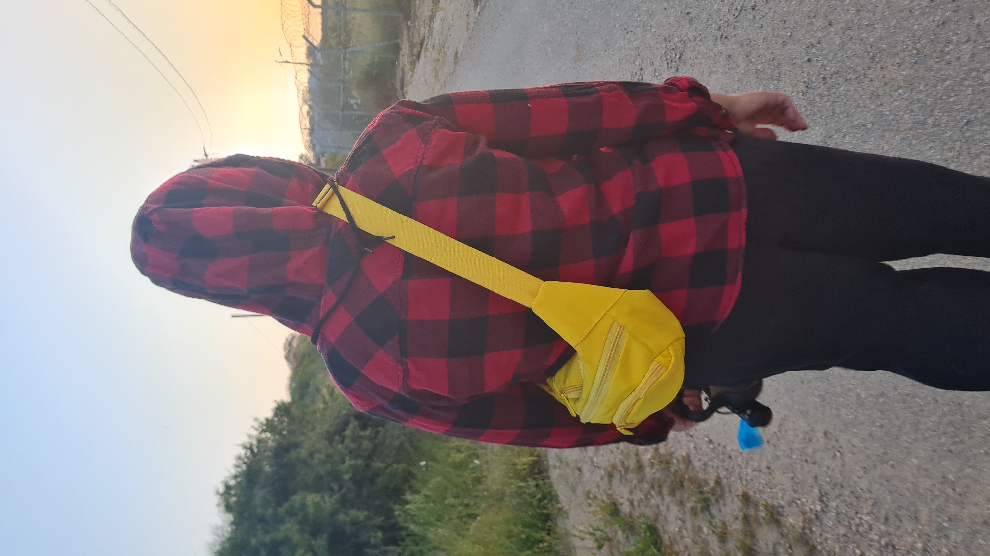 A person wearing a red and black hooded flannel shirt, black pants, and a yellow fanny pack stands on a paved surface. The person's face is obscured by the hood. In the background, there are trees, a fence, and the sky, which is brighter towards the horizon, possibly indicating sunset or sunrise. The location is Lachish Regional Council, Israel.