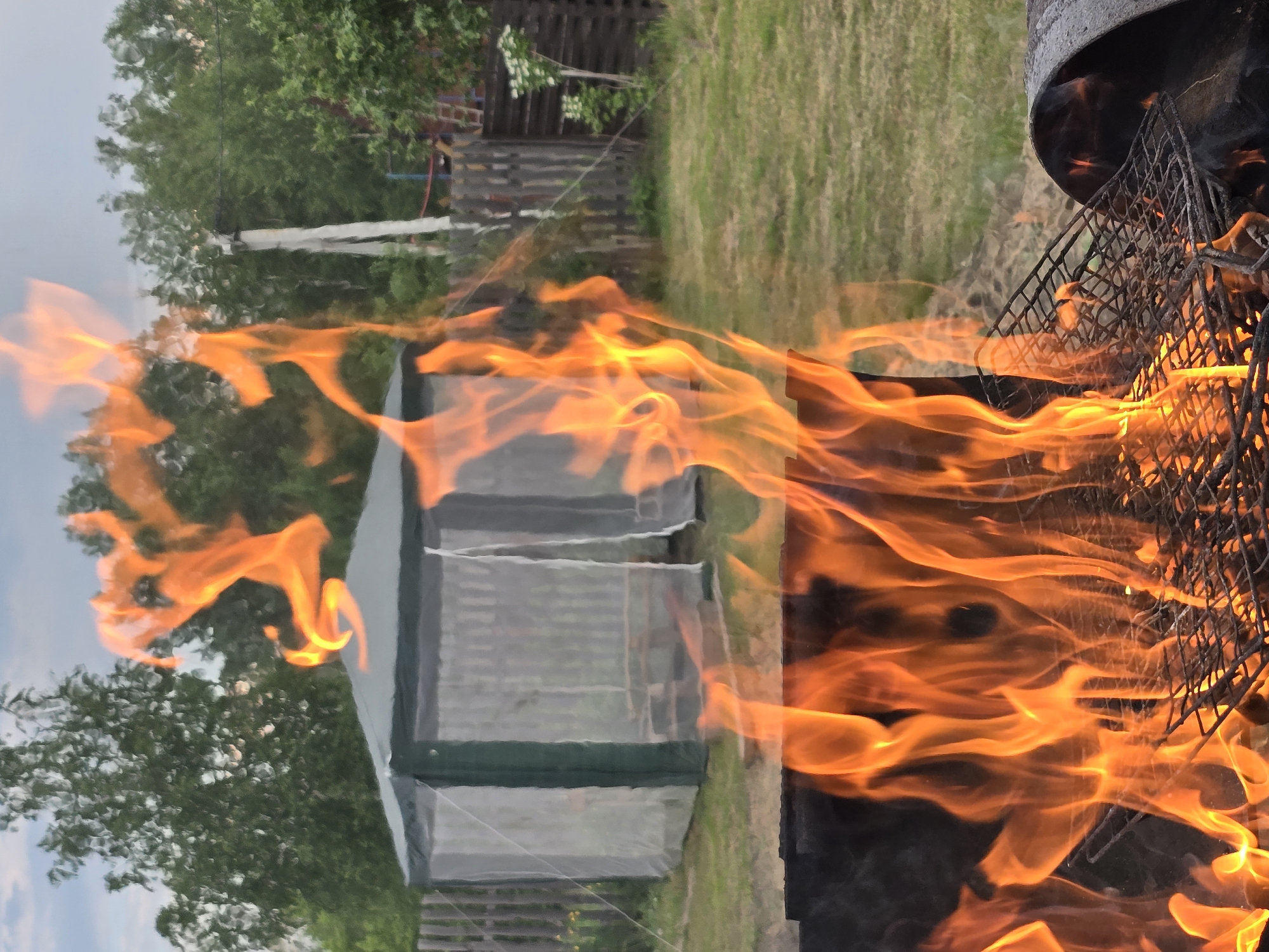 In an outdoor setting, a fire burns in a grill in the foreground, with flames licking upwards and obscuring the view of the grill grate. Behind the fire, a green and white gazebo stands on a grassy lawn. A wooden fence runs along the back of the yard, and trees form a backdrop against a cloudy sky. The scene appears to be a backyard or a park.