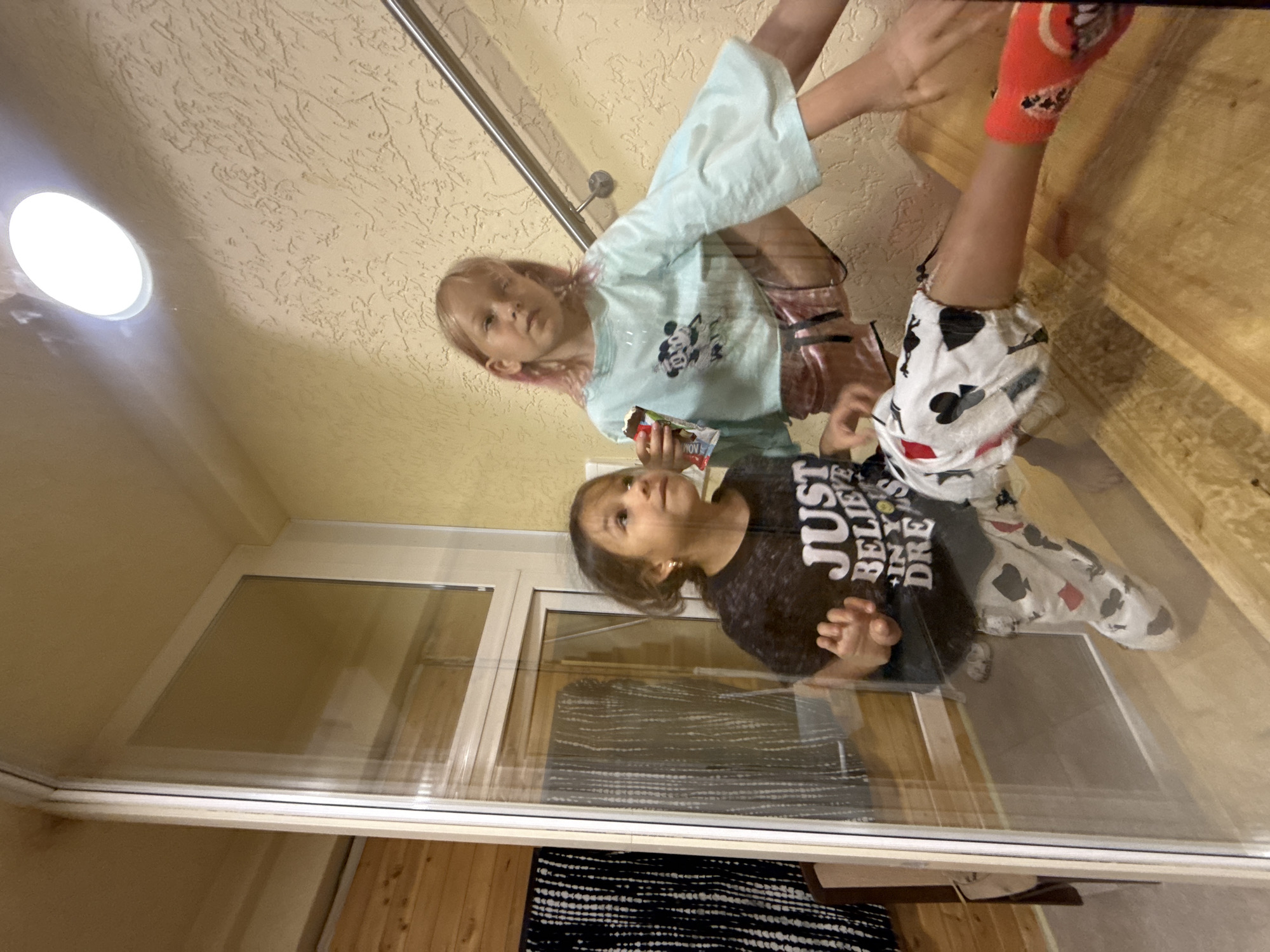 In the city of Unknown, Unknown, two young girls are positioned on wooden stairs indoors. The girl on the left, with dark hair, wears a black t-shirt with white lettering that reads "JUST BELIEVE IN YOUR DREAMS" and white pants with a pattern of black and red spade and club symbols. She is looking to the right and has her left hand raised. The girl on the right, with blonde hair and pink streaks, wears a light blue, bell-sleeved t-shirt with a Mickey Mouse graphic and pink shorts. She is holding a chocolate bar in her right hand and looking forward. A metal handrail is visible to the right of the girls, and a circular light fixture is present on the yellow, textured ceiling above. The scene is viewed through a glass surface, creating reflections.