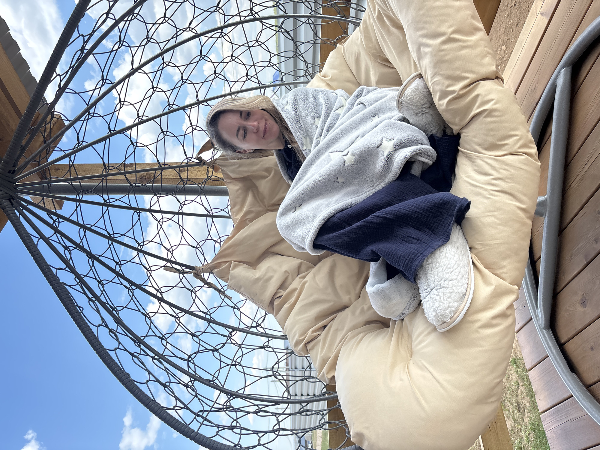 A person is reclined in a beige padded hanging chair with a gray metal frame, likely in Chaikovsky, Russia. The individual is wrapped in a light gray blanket with white star patterns and a navy blue covering, and is wearing white fuzzy slippers. The chair is positioned on a wooden deck with grass visible underneath, and the background shows a blue sky with white clouds visible through the chair's frame.