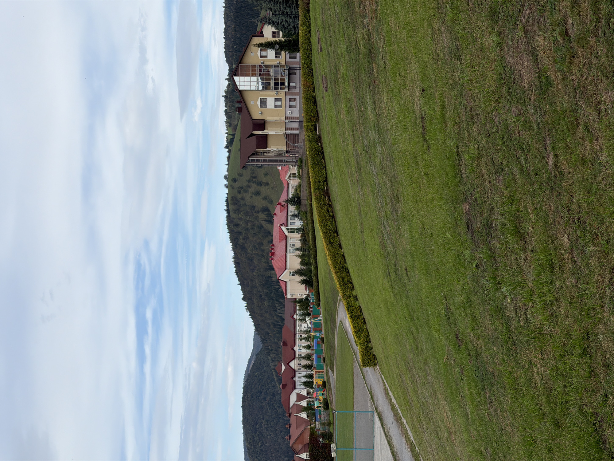 The image shows a cluster of buildings with red roofs, a playground, and a sports field on a grassy hill. In the background, a larger building with a yellow facade and a modern glass-covered section stands out. The entire scene is nestled against a backdrop of lush green hills and a cloudy sky. The location is identified as the city of Assy, Russia, and the image depicts a resort or residential area. There are no people visible in the image, and the scene appears to be tranquil and undisturbed. The weather is overcast.