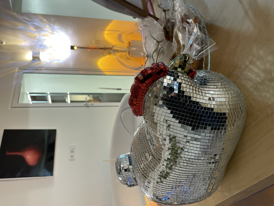 A disco ball sculpture resembling a chicken sits on a light brown surface. The chicken has a red mosaic comb and a mosaic tail. The body consists of small, reflective silver tiles. In the background is a white wall with a dark framed picture and a white window. Overhead lighting casts a warm glow on the scene. Packaging materials are visible behind the chicken, suggesting it may be newly acquired.