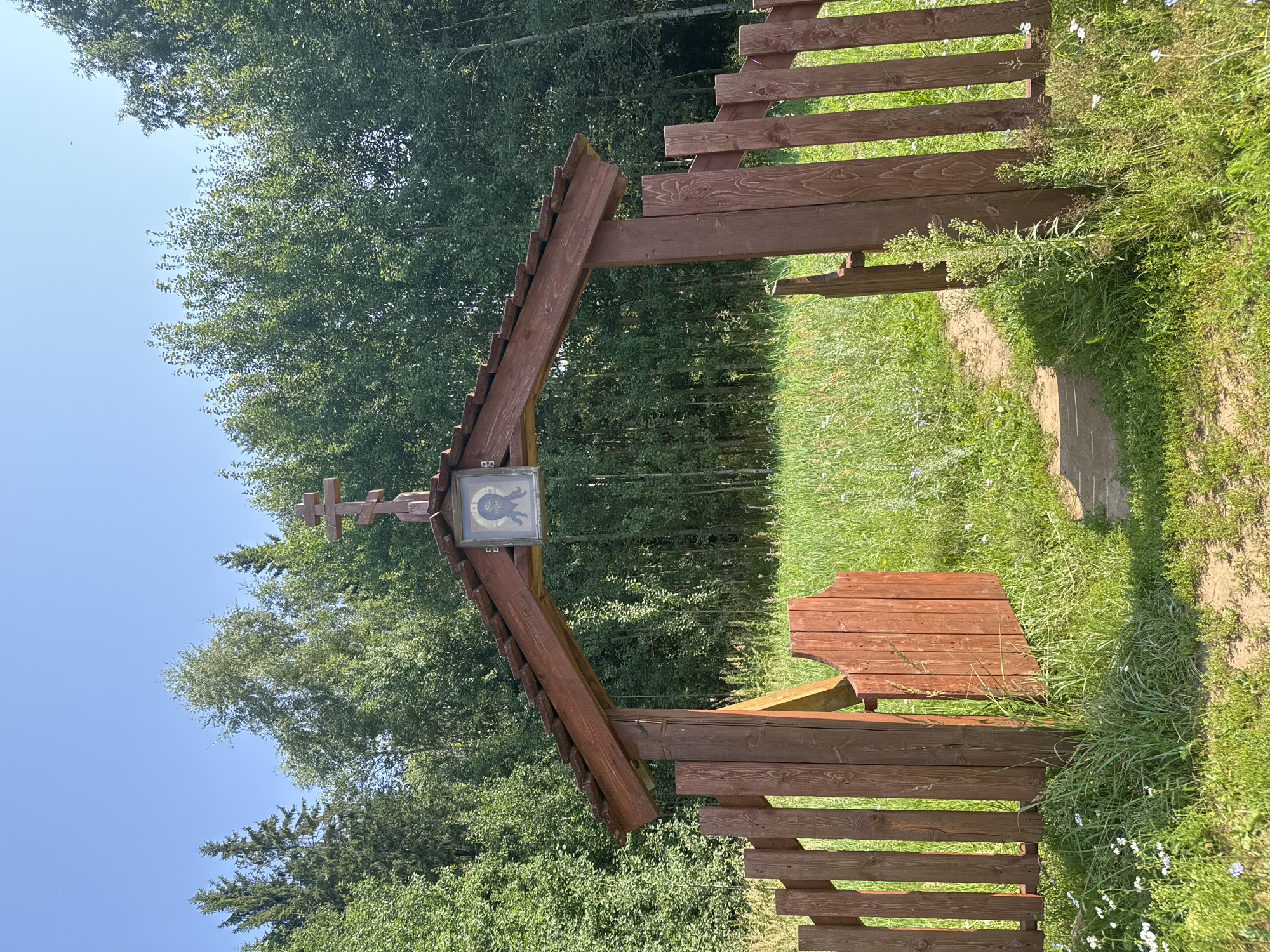 This is a full, eye-level, outdoor shot of a wooden gate and archway leading to a grassy field surrounded by trees. The gate is brown wood with vertical planks and an open section on the left. The archway is also brown wood, with a tiled roof and a cross and an icon of Jesus on the front. The background is filled with a dense forest of green trees under a clear blue sky. The lighting suggests it is daytime, likely midday due to the bright sunlight. No people or animals are visible in the frame. The scene appears to be in a rural or semi-rural location, possibly on the outskirts of a settlement.