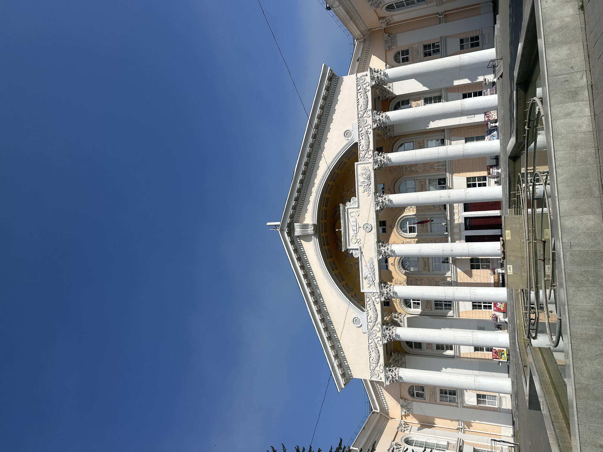 The image shows a wide, low-angle shot of the Biysk Drama Theater in Biysk, Russia. The building is grand, with a pale yellow facade and a prominent portico supported by numerous large white columns. Above the columns, there is an arched area and decorative moldings. Several windows are visible on the facade, some with arched tops. A Russian flag is flying on a pole to the right of the entrance. In the foreground, a shallow, rectangular fountain with curved interior elements is visible, the water reflecting the blue sky and the building. The sky is clear and bright blue. To the left of the building, a dark green pine tree is partially visible. There are posters displayed near the entrance of the building and planters with flowers. The scene appears to be a public square or plaza in front of the theater.