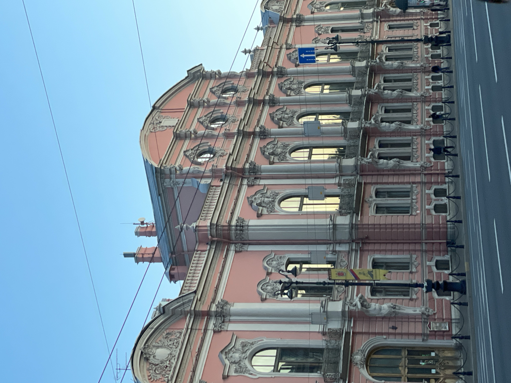 This upward-angled shot captures a majestic, pale pink building with opulent white and light gray architectural details, characteristic of historic European architecture, consistent with Saint Petersburg, Russia. The building features a distinctive curved facade, particularly noticeable towards its upper floors and roofline. Multiple rows of windows, varying from rectangular to arched and circular/oval, are integrated into the elaborate design. Many windows are flanked by decorative columns or pilasters and topped with ornate stucco work, including shell motifs and floral patterns. A prominent feature is the series of sculpted figures, possibly atlantes or caryatids, depicted in dynamic poses as they appear to support the weight of the upper floors and window sills.
The windows reflect a warm, golden light, indicating bright daytime sunshine under a clear blue sky. Thin, dark wires, likely for electrical or public transport infrastructure, traverse the sky in front of the building. On the street level, a paved road with a white lane marking is visible, bordered by decorative black street lamps. A blue rectangular traffic sign with a white arrow pointing straight ahead is mounted on the building's facade. Another yellow sign is seen near a street lamp, but its details are indistinct. The overall impression is one of grand, historic architecture bathed in sunlight.