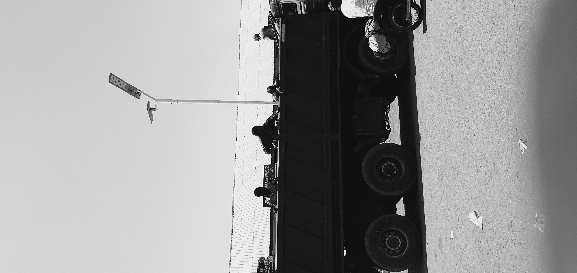 The image depicts a street scene in Bukuru, Nigeria, under a clear sky. A large, dark-colored truck with its bed loaded is the central focus. Several individuals are positioned around and within the truck's bed. One person, wearing a light-colored shirt, leans over the side of the truck bed, appearing to be engaged in an activity. To their right, another individual in a dark jacket also leans over the truck bed. Further to the right, a third person, wearing a cap and a light-colored garment, is seen standing behind the truck. In the foreground, a motorcycle is parked, with a person seated on it wearing a light-colored shirt. The ground is asphalt, with some scattered debris. A street lamp with solar panels is visible in the background, to the left of the truck. A building with a corrugated metal roof is situated behind the truck and the people. Signs are also visible on the building, one with the text "FIT WEARS". The image is rendered in black and white.