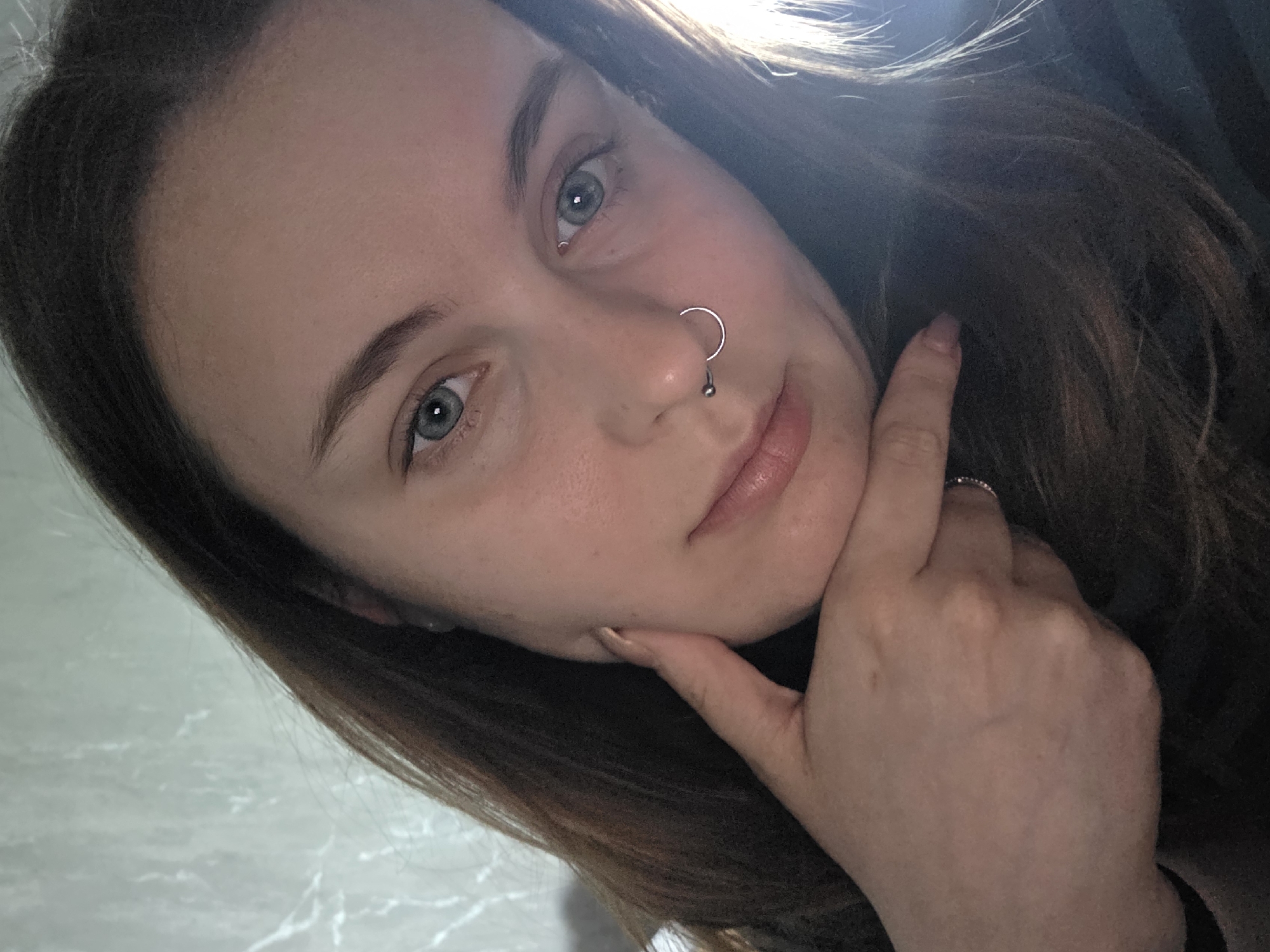 The image is a close-up, frontal selfie of a young woman with long, brown hair. She has light skin, blue-grey eyes, and a septum piercing. Her left hand is resting on her chin, and she is wearing a ring on her left index finger. The background appears to be a wall with a marbled pattern. There are no visible landmarks that can definitively place this image in Minsk, Belarus.