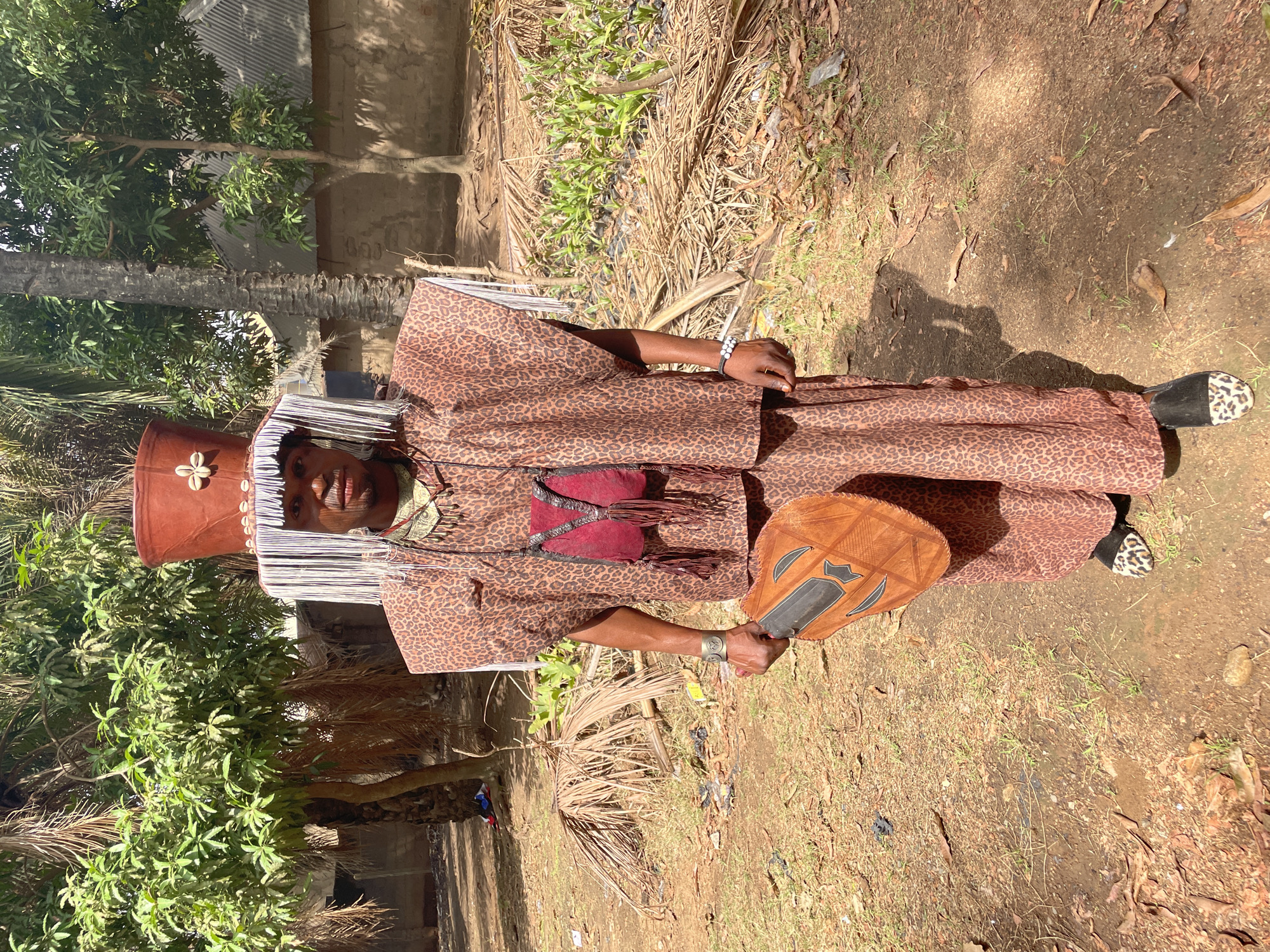 Akwanga, Nigeria: A person in traditional attire stands on a grassy, slightly dusty ground, surrounded by sparse vegetation and trees. The individual wears a brown leopard-print outfit, a tall brown hat adorned with shells, and white beaded strands hanging from the hat's brim. They hold a brown fan in their right hand and wear a bracelet on their left wrist. A maroon bag hangs from their shoulder. One shoe has a black and white leopard-print pattern. A building and more trees are visible in the background.