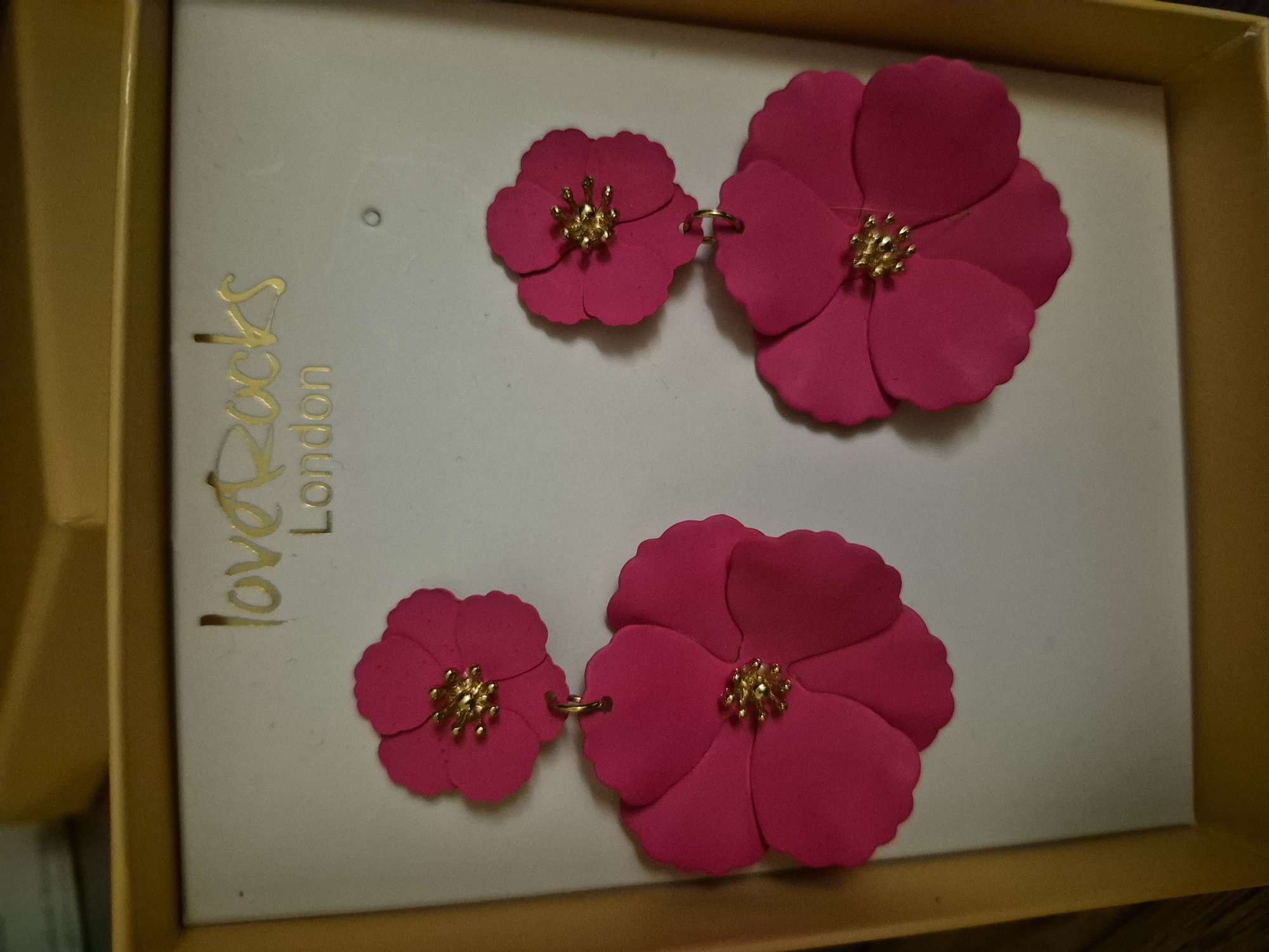 The image shows a pair of bright pink flower-shaped drop earrings with gold centers, displayed in a white cardboard box with the brand name "love Rocks London" printed in gold. The earrings are the primary focus, with a smaller flower at the top and a larger one dangling below. The box is slightly angled, revealing a yellow border. The background is blurry and out of focus.