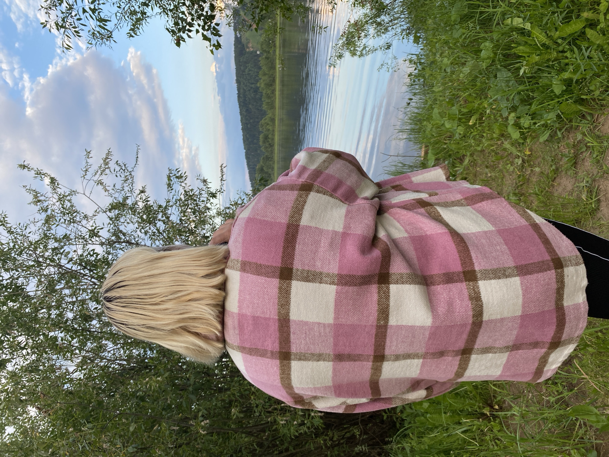 The image shows a young woman with blonde hair, seen from behind, wearing a pink and cream plaid shirt jacket over black leggings. She is standing on a grassy area next to a calm body of water, likely a river or lake, with a treed hillside in the background. The sky is partly cloudy with shades of blue, white, and hints of pink and orange from the setting sun. The location is identified as Noviy, Russia.