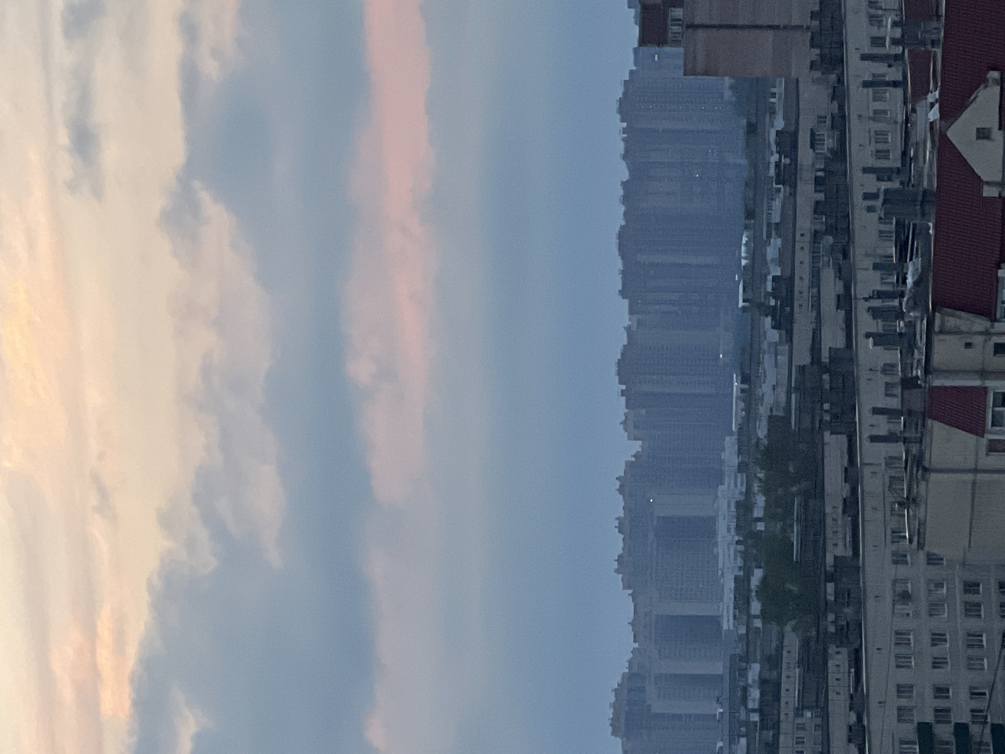 The image shows a cityscape at dusk. The sky is filled with soft, diffused clouds in hues of pink, orange, and blue, suggesting either sunrise or sunset. In the middle ground, a dense cluster of high-rise apartment buildings stretches across the horizon, appearing as silhouettes against the atmospheric sky. The buildings are uniform in their architecture, with many floors and similar window patterns. In the foreground, the rooftops of lower buildings are visible, some featuring the characteristic grey facades and visible ventilation pipes of Soviet-era housing blocks. A few green trees are visible amidst these lower structures, adding a touch of nature to the urban landscape. The overall impression is of a large residential area within a Russian city.