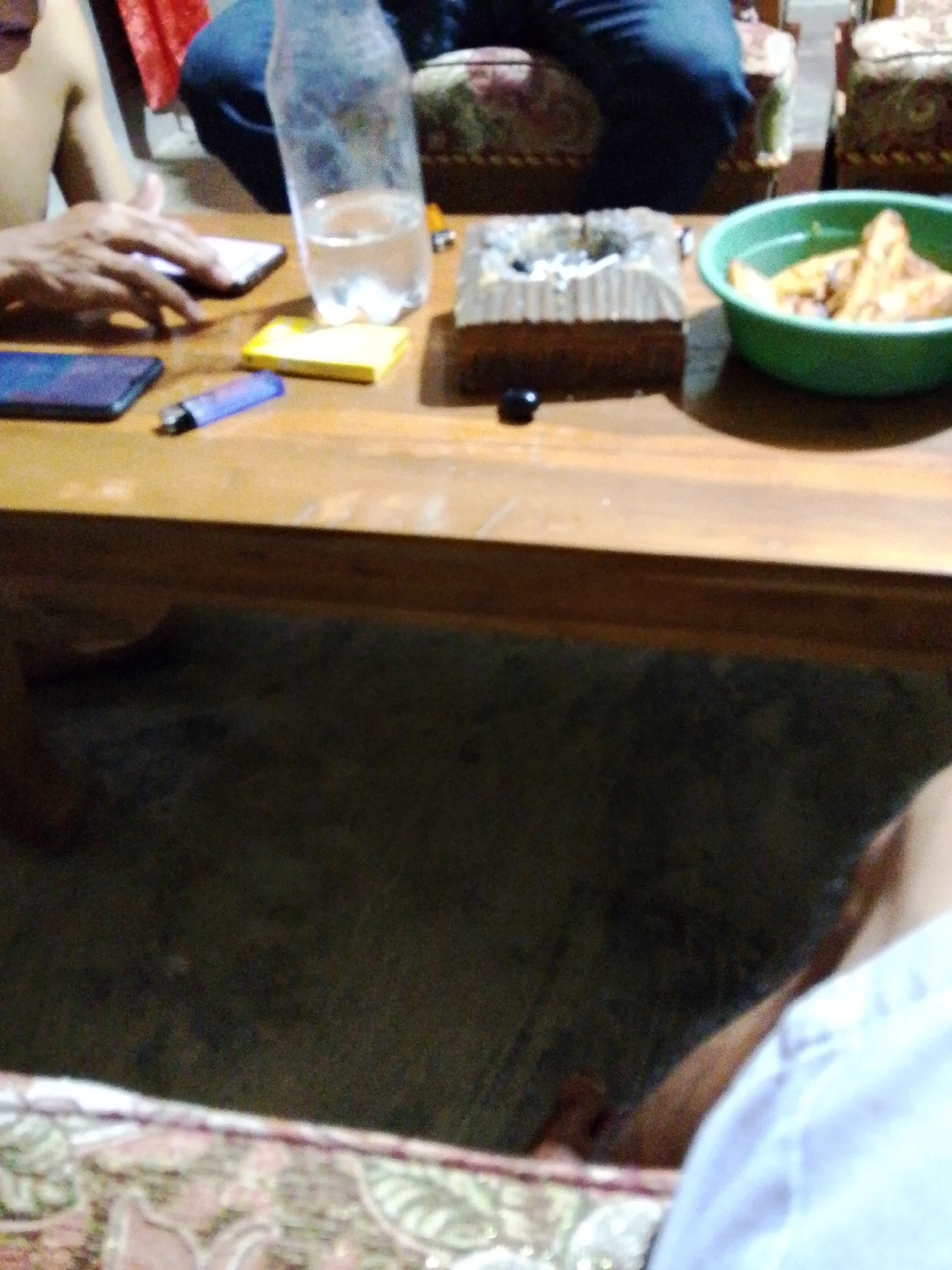 A low-angle, slightly out-of-focus indoor shot captures a wooden table in Madukoro, Indonesia. On the table, a clear plastic bottle filled with clear liquid sits next to a square, multi-colored ashtray containing cigarette butts and ash. To the right of the ashtray is a green bowl filled with fried food, possibly chicken or potatoes. In the foreground, a blue lighter lies on the table near a yellow object, possibly a pack of cigarettes or gum. A hand, appearing to be reaching for a dark-colored smartphone, is visible on the left side of the frame. In the background, a person in blue jeans is seated, and a portion of a red fabric can be seen draped. The overall lighting is dim, and the scene suggests a casual gathering.