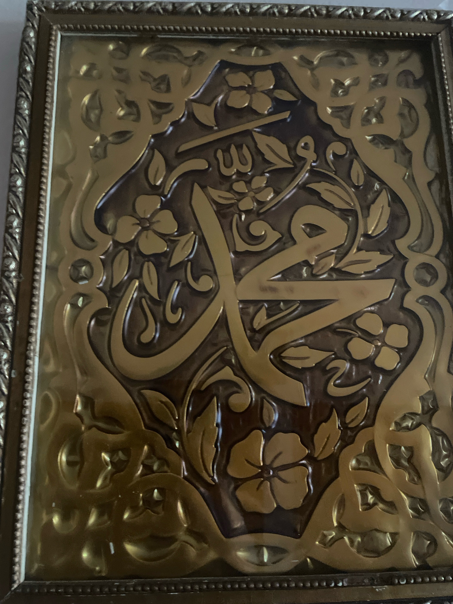 The image shows a close-up of a framed piece of art featuring Islamic calligraphy. The calligraphy, rendered in gold-tone metal, appears to spell out the name of the Prophet Muhammad. The calligraphy is set against a dark background and surrounded by an ornate, embossed floral and scrollwork pattern, also in a gold-tone metal. The frame itself is metallic with a textured detail. The overall impression is of a religious or decorative artifact. The specific location in Ghazaouet, Algeria, is not identifiable from the image itself, as it is a close-up of the artwork and lacks any contextual environmental clues of the city.
