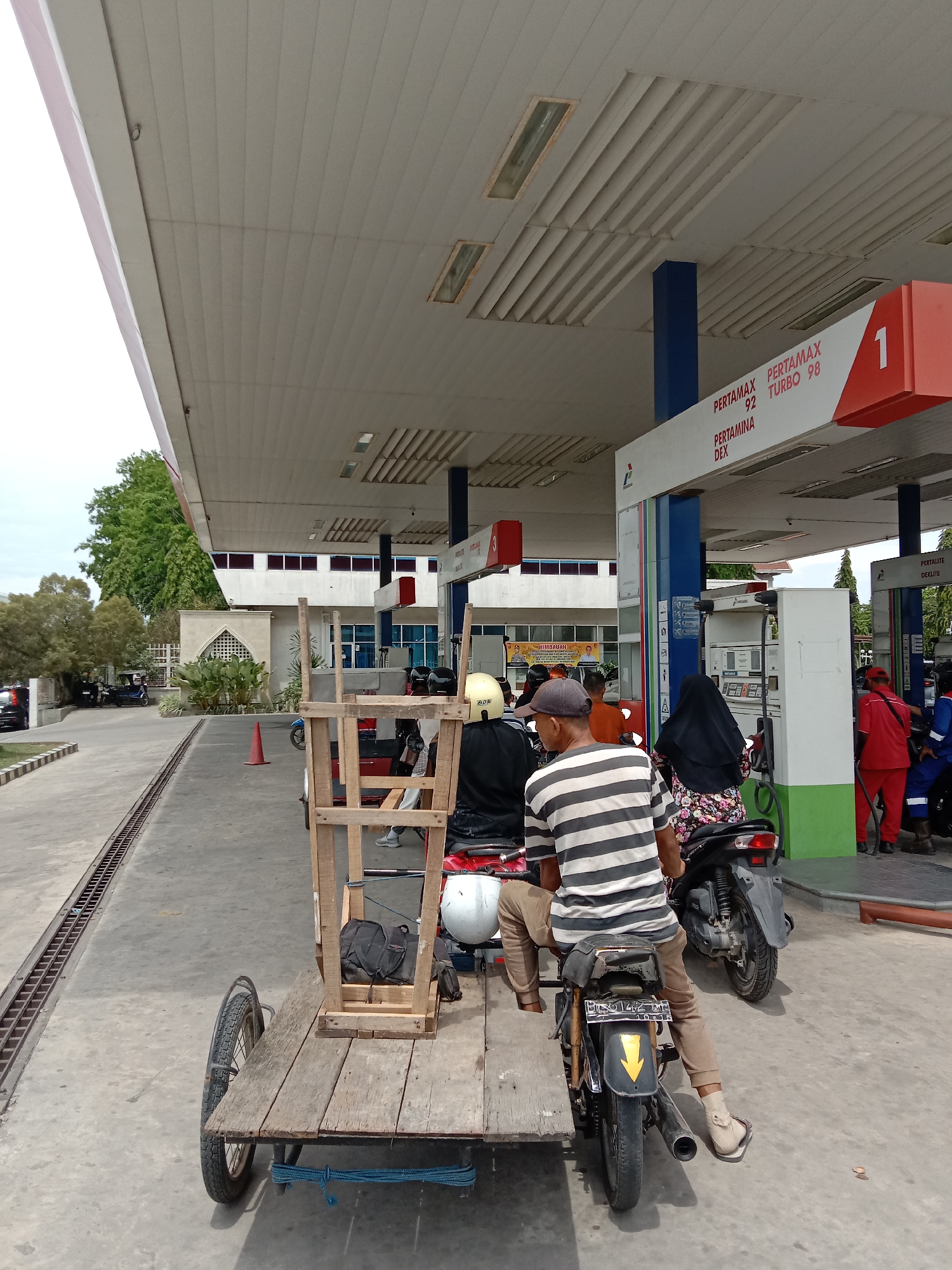 The image shows a scene at a Pertamina gas station in Lhokseumawe, Indonesia. Several motorbikes are lined up at a pump. The first motorbike has a wooden cart attached to it. A man wearing a striped shirt is sitting on the motorbike. Behind him is a motorbike ridden by a woman wearing a black hijab. Two attendants in red uniforms are serving at the gas pump. The gas station has a large canopy, and the sign above the pump indicates the available gas types: Pertamax 92, Pertamax Turbo 98, and Pertamina Dex. In the background, there is a building with windows and trees.